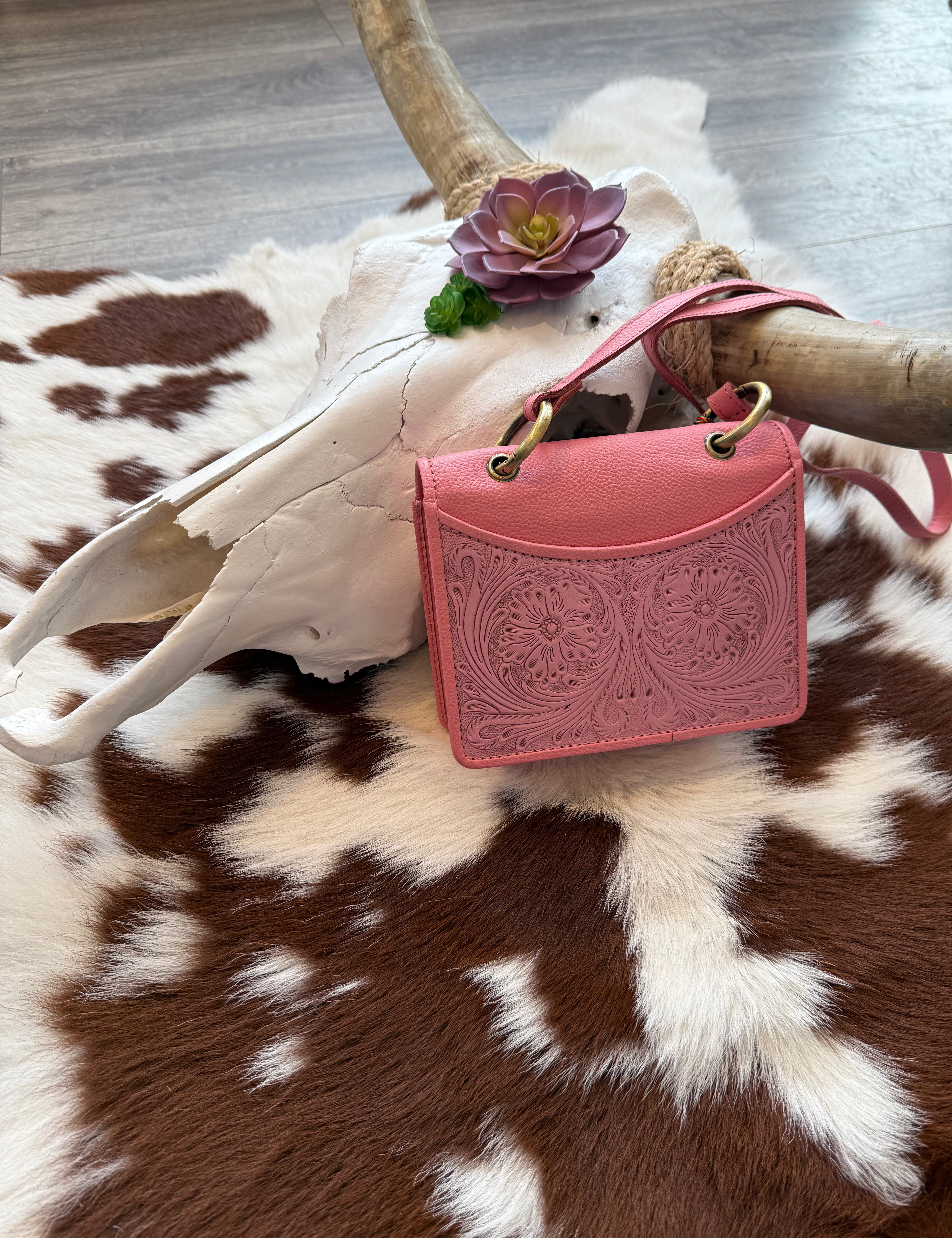 A pink embossed leather handbag with intricate floral tooling rests against a white cow skull adorned with artificial succulents. The bag has gold-toned metal rings attaching the handle and a matching pink crossbody strap. The background features a cowhide rug with brown and white patches, complementing the rustic, Western-inspired aesthetic