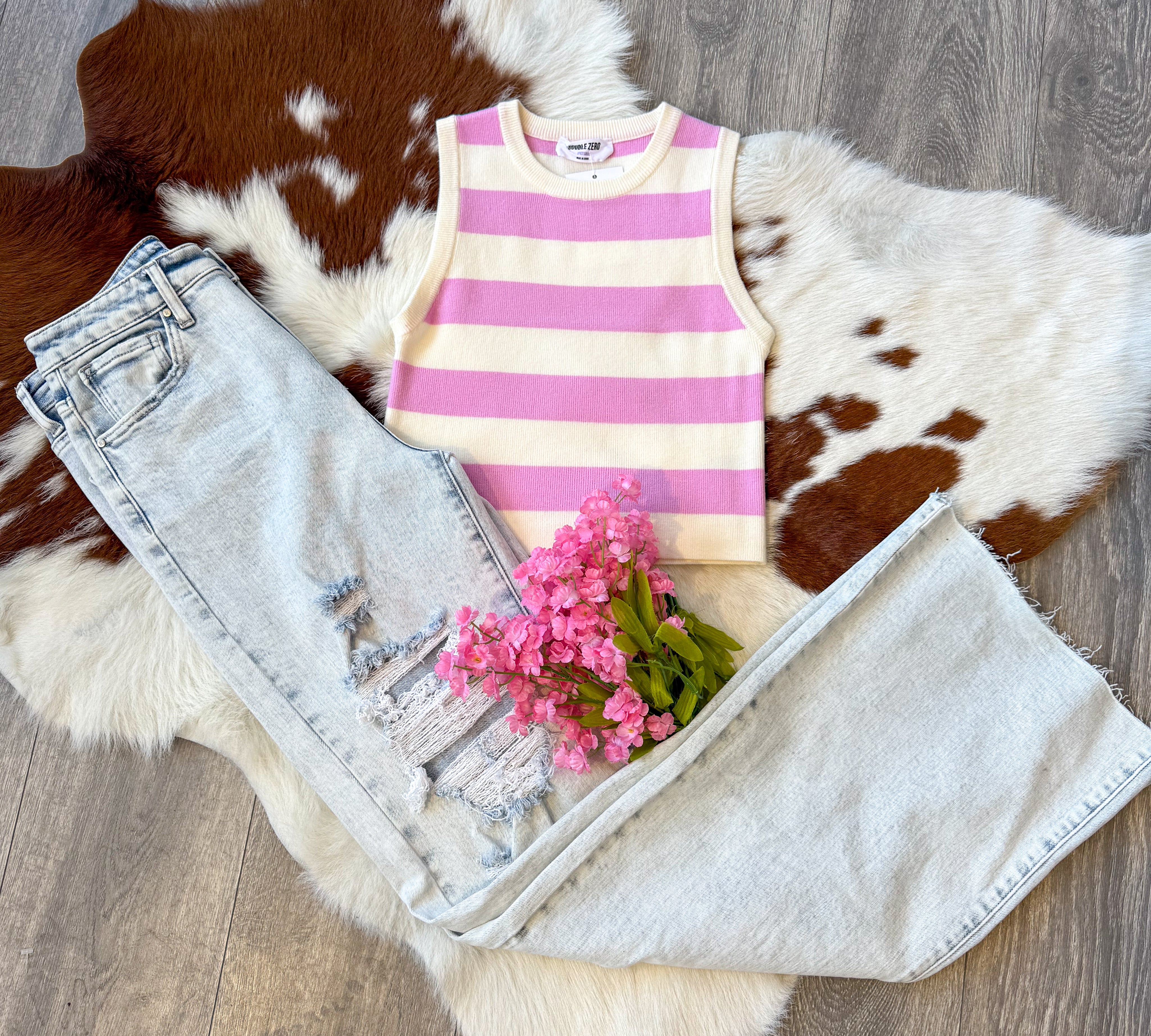 A trendy outfit laid flat on a brown and white cowhide rug, featuring a sleeveless pink and white striped knit tank top paired with light-wash distressed wide-leg jeans. The jeans have ripped details on one leg, adding a casual, edgy touch. A small bouquet of pink flowers is placed on top of the jeans, enhancing the feminine aesthetic. The display is set on a wood floor, complementing the rustic vibe