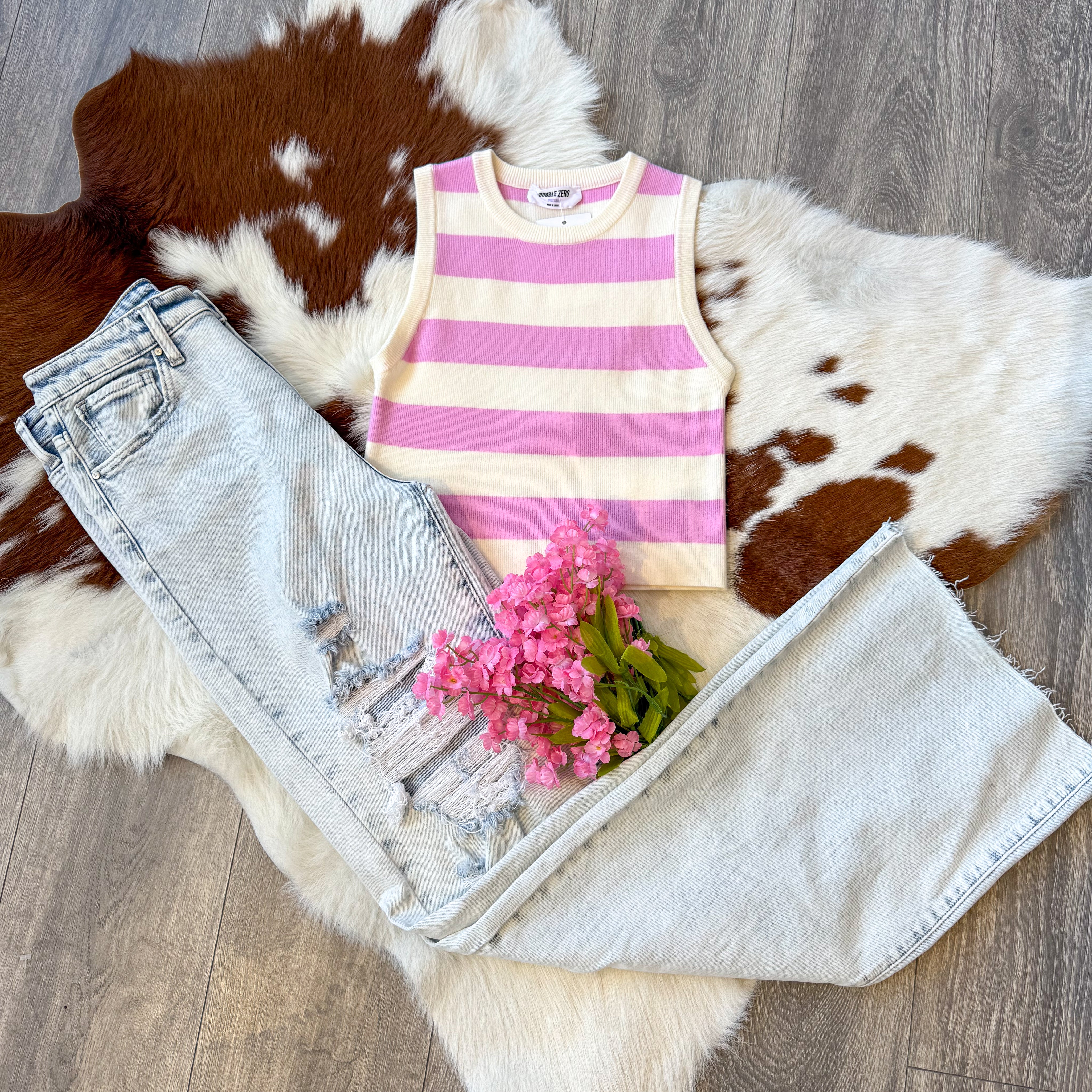 A trendy outfit laid flat on a brown and white cowhide rug, featuring a sleeveless pink and white striped knit tank top paired with light-wash distressed wide-leg jeans. The jeans have ripped details on one leg, adding a casual, edgy touch. A small bouquet of pink flowers is placed on top of the jeans, enhancing the feminine aesthetic. The display is set on a wood floor, complementing the rustic vibe