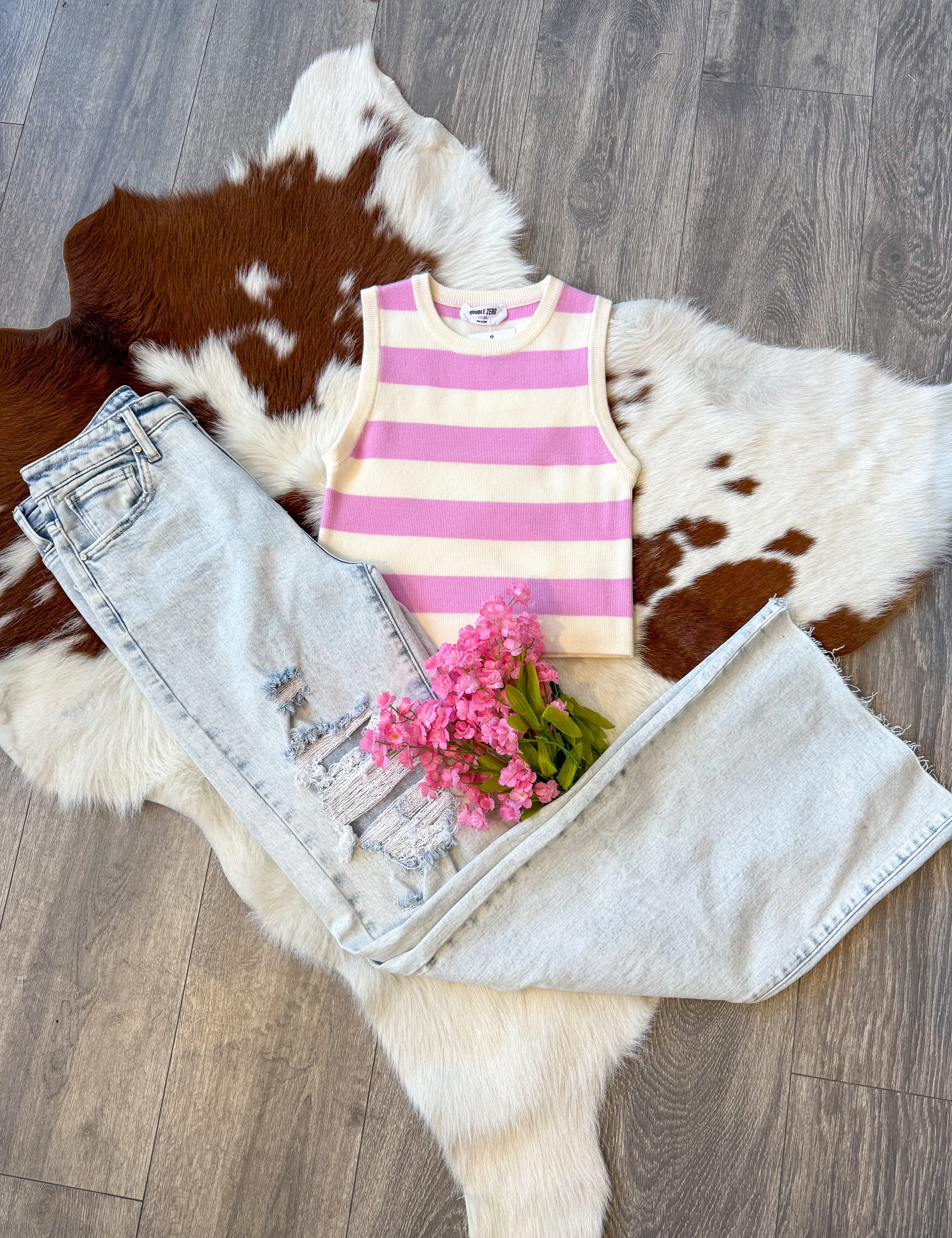 A trendy outfit laid flat on a brown and white cowhide rug, featuring a sleeveless pink and white striped knit tank top paired with light-wash distressed wide-leg jeans. The jeans have ripped details on one leg, adding a casual, edgy touch. A small bouquet of pink flowers is placed on top of the jeans, enhancing the feminine aesthetic. The display is set on a wood floor, complementing the rustic vibe