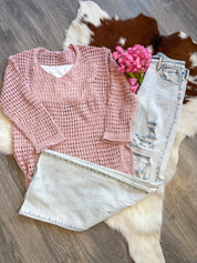 A cozy yet stylish outfit laid flat on a brown and white cowhide rug, featuring a soft pink open-knit sweater layered over a white tank top, paired with light-wash distressed wide-leg jeans. The sweater has a relaxed fit and a breathable texture, perfect for layering. A bouquet of pink flowers adds a charming, feminine touch to the display, which is set on a wood floor for a rustic-chic vibe.