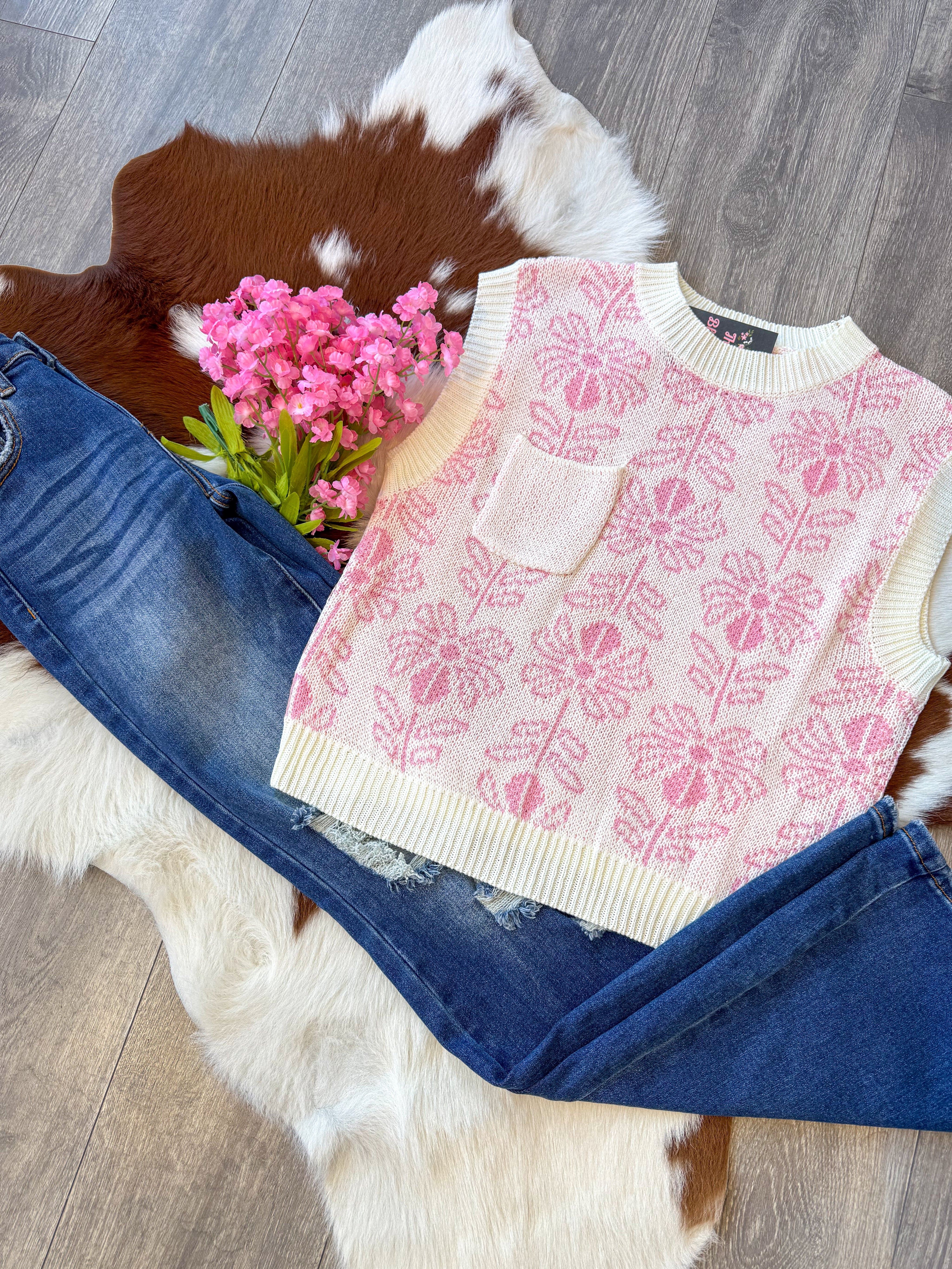 A cute and cozy outfit laid flat on a brown and white cowhide rug, featuring a cream and pink floral knit sweater vest with a small front pocket. The vest is paired with dark-wash distressed jeans for a trendy yet effortless look. A bouquet of pink flowers adds a charming touch, while the wood floor enhances the rustic-chic vibe.