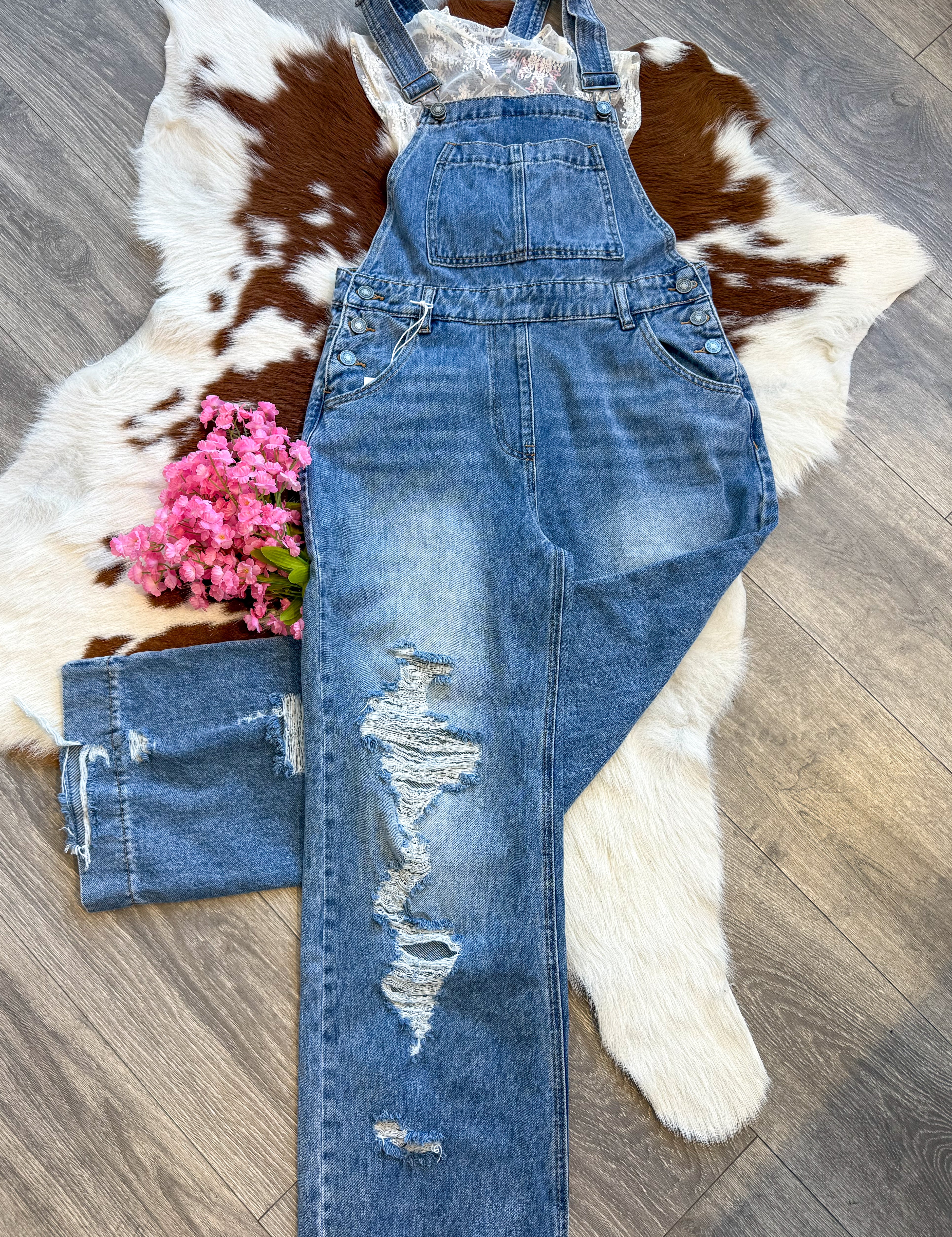 A stylish distressed denim overall laid flat on a brown and white cowhide rug. The overalls feature a front bib pocket, adjustable straps, and ripped details on one leg. A delicate white lace top peeks out from underneath, adding a feminine touch. A small bouquet of pink flowers is placed beside the outfit for a charming accent. The display is set on a wood floor, enhancing the rustic aesthetic.