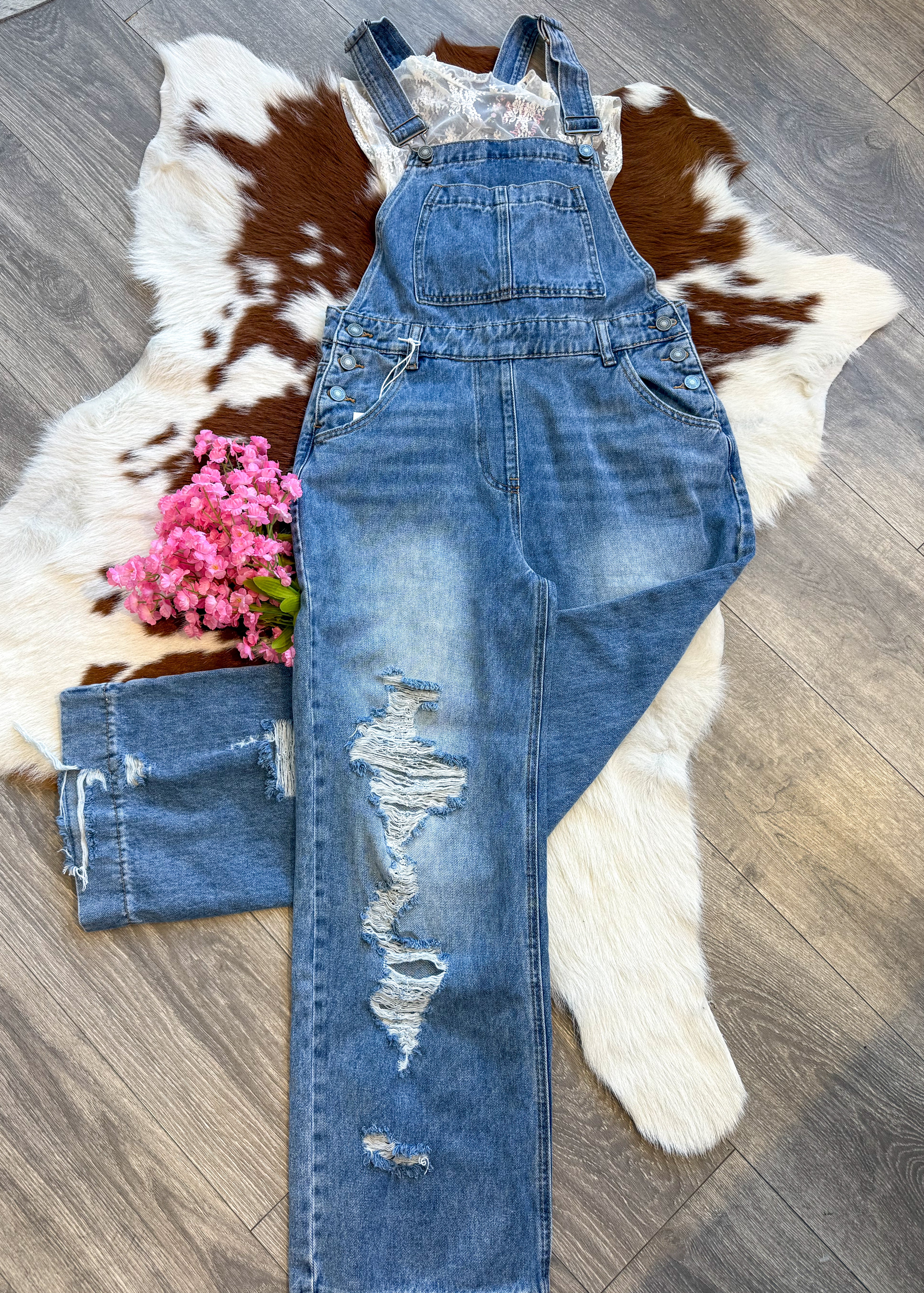 A stylish distressed denim overall laid flat on a brown and white cowhide rug. The overalls feature a front bib pocket, adjustable straps, and ripped details on one leg. A delicate white lace top peeks out from underneath, adding a feminine touch. A small bouquet of pink flowers is placed beside the outfit for a charming accent. The display is set on a wood floor, enhancing the rustic aesthetic.