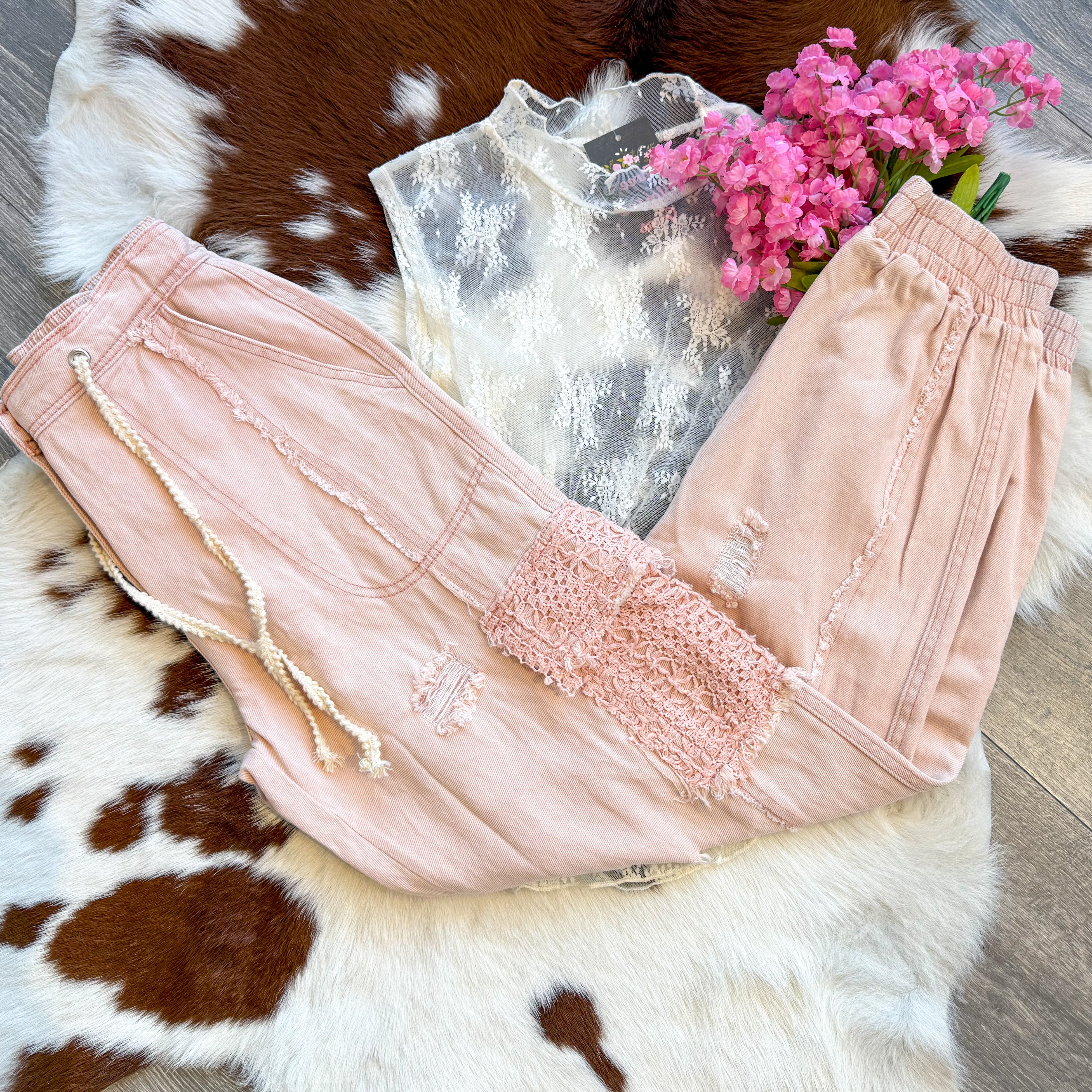 A soft and feminine outfit laid flat on a brown and white cowhide rug, featuring blush pink distressed jogger pants with lace patch details and a drawstring waist. The pants are paired with a sheer white lace sleeveless top with delicate floral embroidery for a romantic touch. A bouquet of pink flowers adds a dreamy, whimsical vibe, while the wood floor enhances the rustic-chic aesthetic