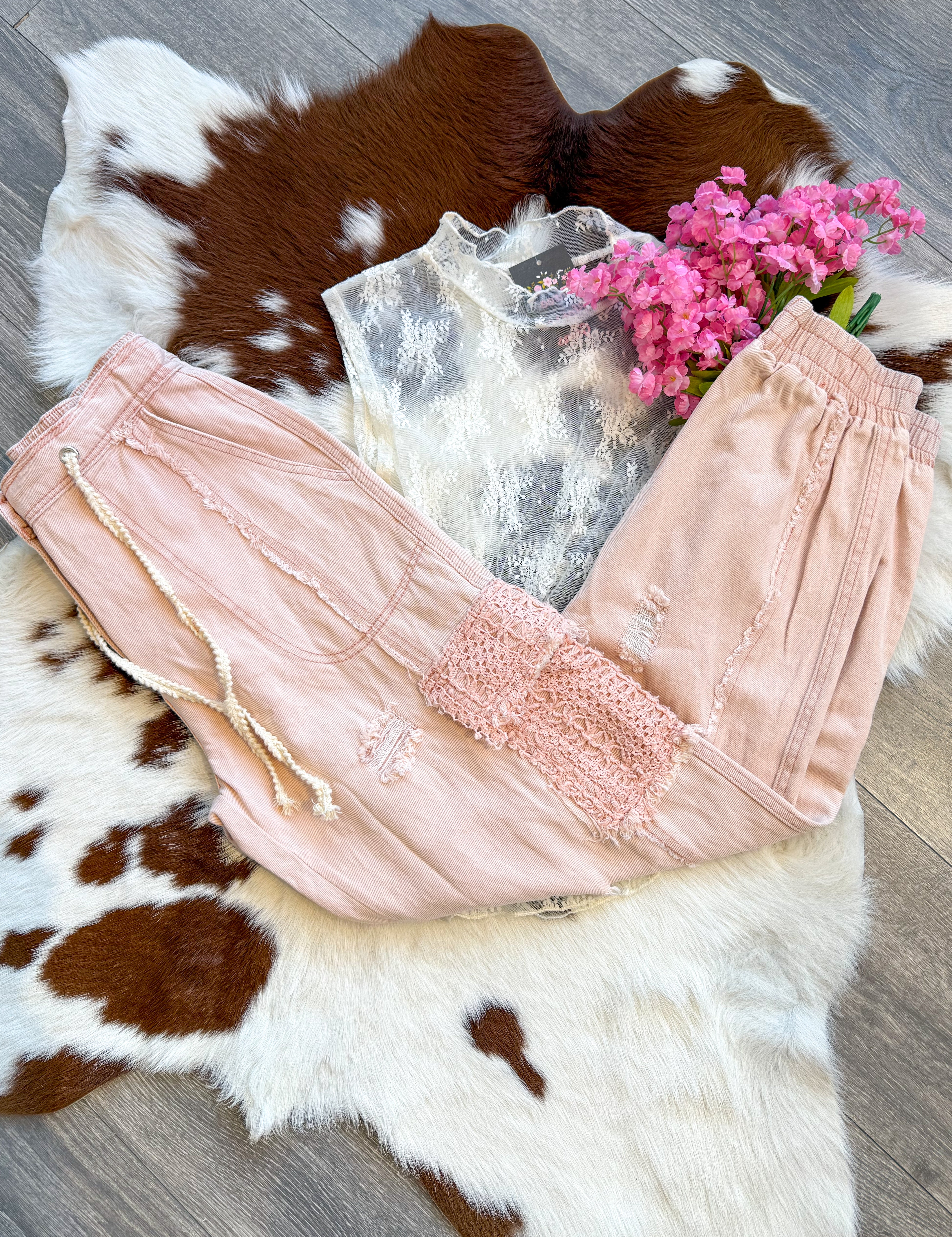 A soft and feminine outfit laid flat on a brown and white cowhide rug, featuring blush pink distressed jogger pants with lace patch details and a drawstring waist. The pants are paired with a sheer white lace sleeveless top with delicate floral embroidery for a romantic touch. A bouquet of pink flowers adds a dreamy, whimsical vibe, while the wood floor enhances the rustic-chic aesthetic