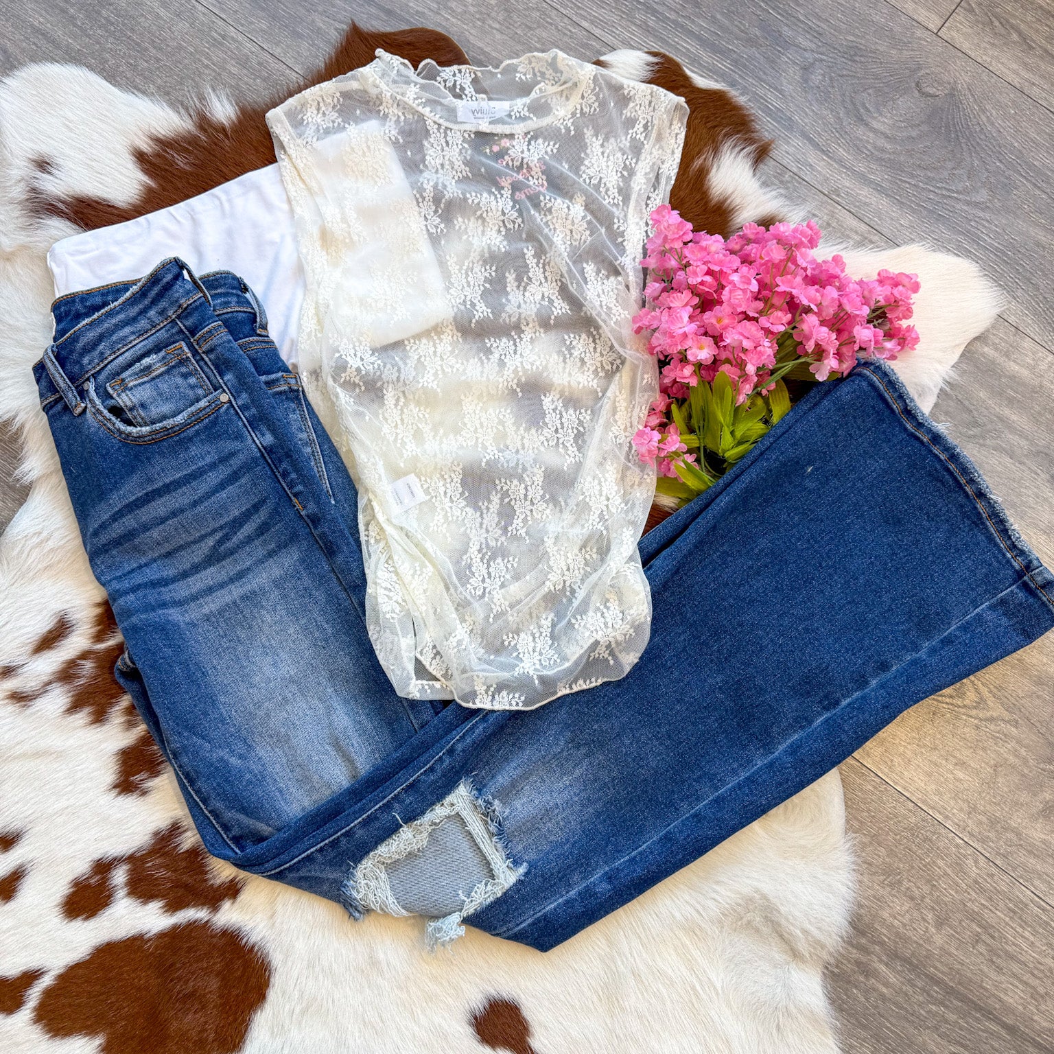 A chic and romantic outfit laid flat on a brown and white cowhide rug, featuring a delicate sheer lace top with floral embroidery layered over a classic white tank. The top is paired with dark-wash distressed jeans for a perfect mix of edgy and feminine. A bouquet of pink flowers adds a soft, whimsical touch, while the wood floor enhances the rustic aesthetic