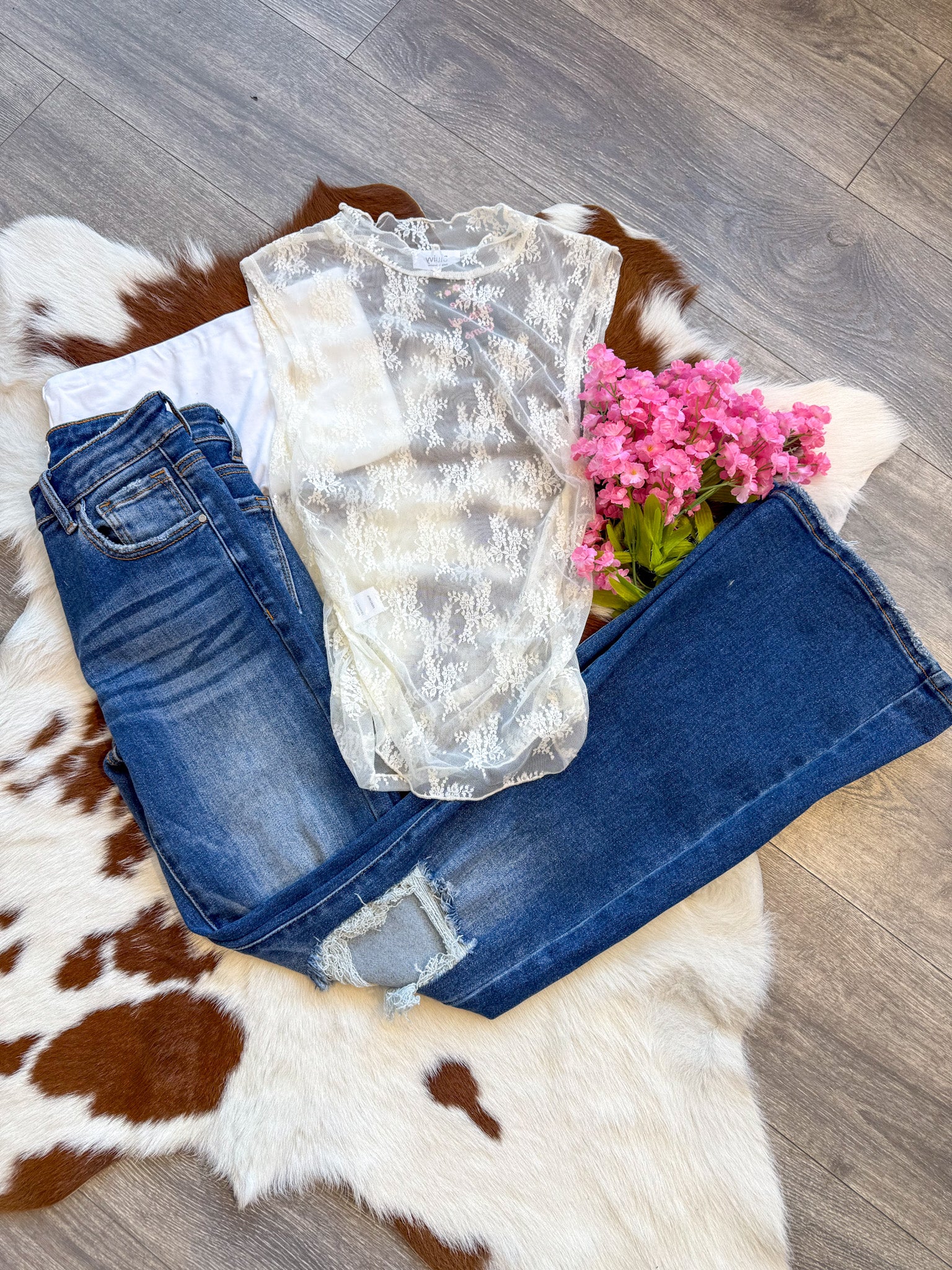 A chic and romantic outfit laid flat on a brown and white cowhide rug, featuring a delicate sheer lace top with floral embroidery layered over a classic white tank. The top is paired with dark-wash distressed jeans for a perfect mix of edgy and feminine. A bouquet of pink flowers adds a soft, whimsical touch, while the wood floor enhances the rustic aesthetic