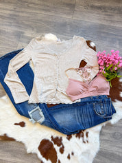 A feminine and flirty outfit laid flat on a brown and white cowhide rug, featuring a beige ribbed long-sleeve top with delicate lace trim, paired with dark-wash distressed jeans. A blush pink bralette with floral embroidery adds a romantic touch, while a bouquet of pink flowers enhances the soft, dreamy aesthetic. The display is set on a wood floor, blending rustic charm with effortless style