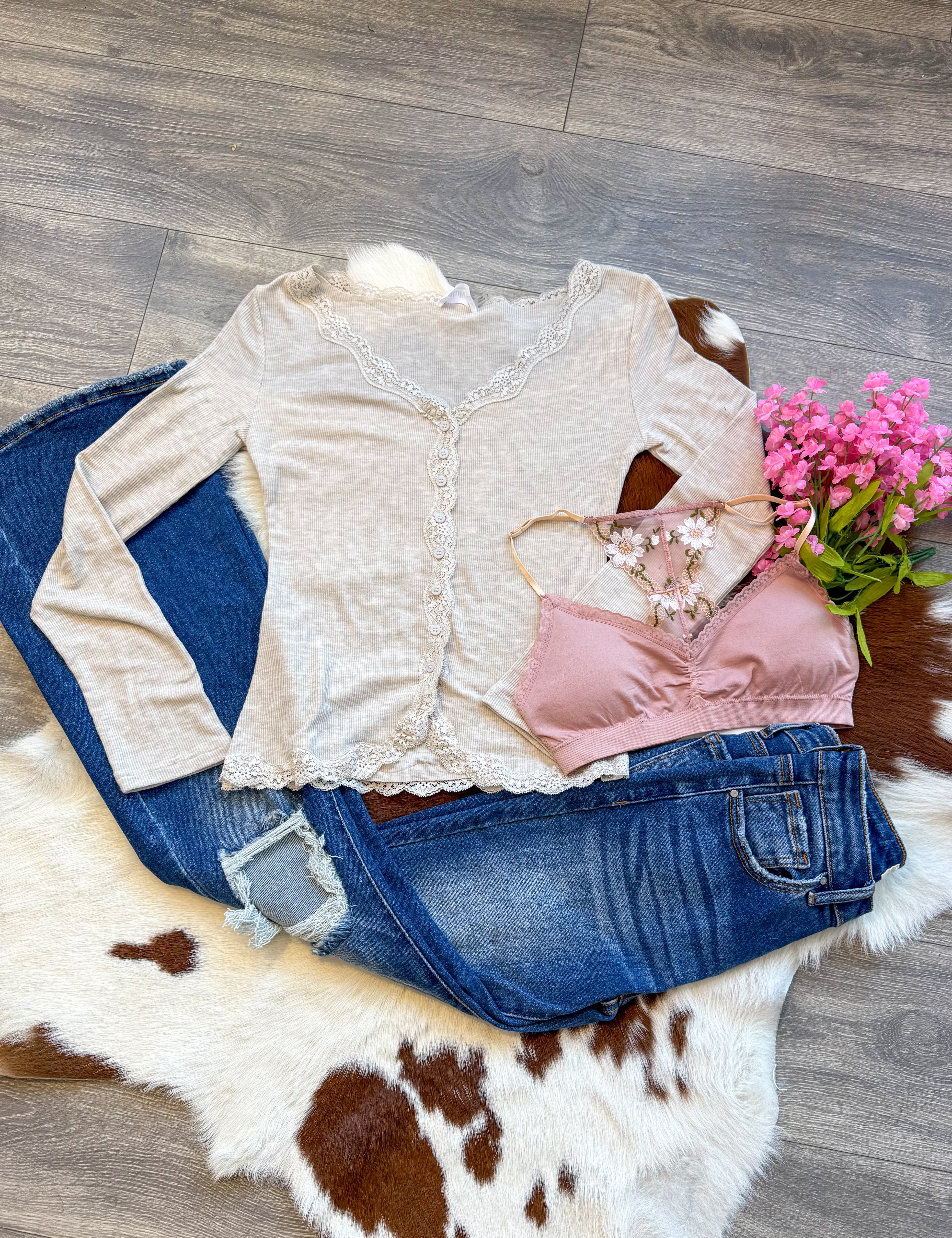 A feminine and flirty outfit laid flat on a brown and white cowhide rug, featuring a beige ribbed long-sleeve top with delicate lace trim, paired with dark-wash distressed jeans. A blush pink bralette with floral embroidery adds a romantic touch, while a bouquet of pink flowers enhances the soft, dreamy aesthetic. The display is set on a wood floor, blending rustic charm with effortless style