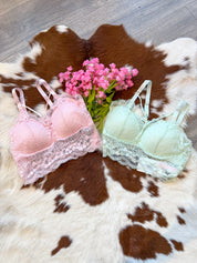 A dreamy and feminine lingerie display featuring two delicate lace bralettes, one in soft pink and the other in pastel mint green, laid flat on a brown and white cowhide rug. Each bralette has intricate lace detailing, adjustable straps, and a scalloped lace trim for a romantic touch. A bouquet of pink flowers adds an extra dose of charm, while the wood floor enhances the rustic-chic aesthetic