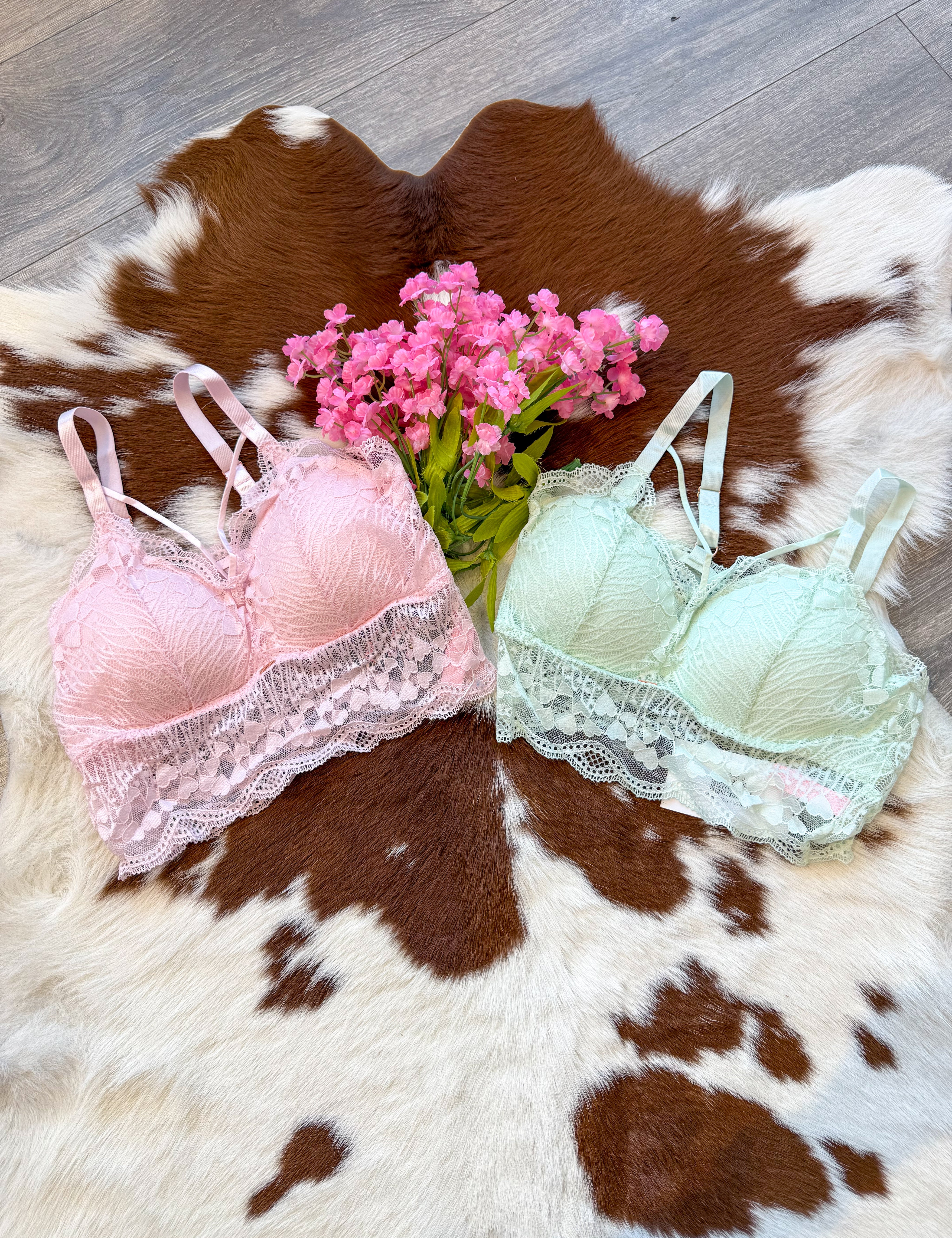 A dreamy and feminine lingerie display featuring two delicate lace bralettes, one in soft pink and the other in pastel mint green, laid flat on a brown and white cowhide rug. Each bralette has intricate lace detailing, adjustable straps, and a scalloped lace trim for a romantic touch. A bouquet of pink flowers adds an extra dose of charm, while the wood floor enhances the rustic-chic aesthetic