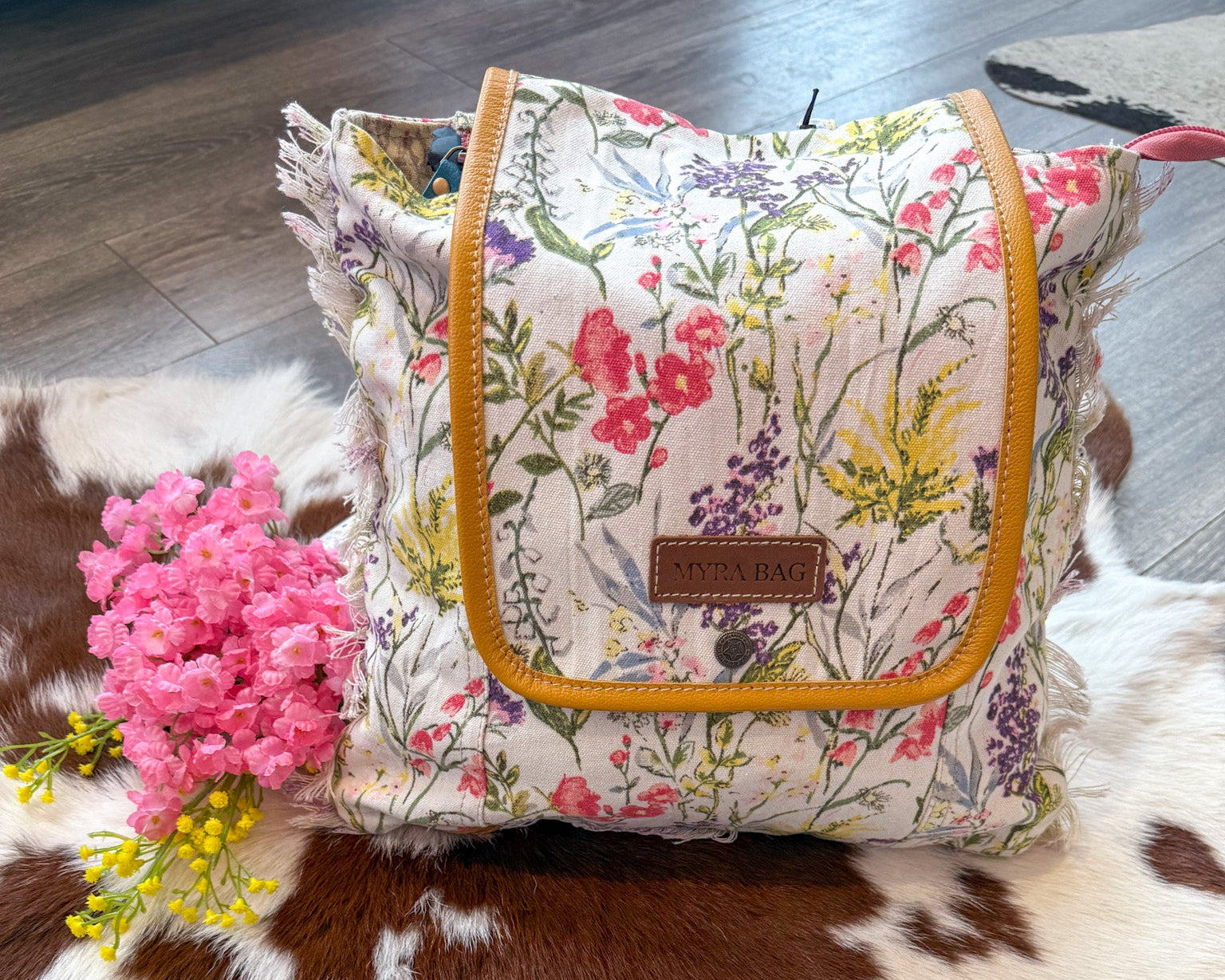 A canvas backpack with a floral meadow print, yellow trim, and a leather accent, displayed on a wooden surface next to pink flowers.