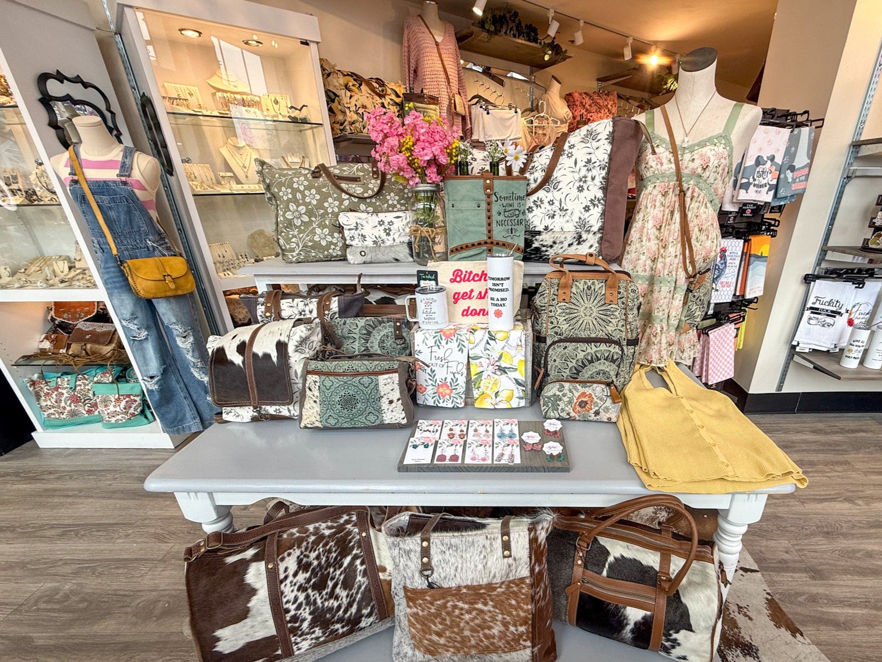 A boutique display featuring a collection of stylish handbags, accessories, and clothing. The table showcases cowhide and patterned bags in various sizes, along with small accessories and a sign that reads, "Bitch, get it done." Behind the table, mannequins are dressed in trendy outfits, including distressed overalls with a striped top and a floral dress. Shelves in the background display jewelry, drinkware, and other boutique items, creating a cozy and fashionable shopping experience. The decor includes so