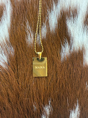 A gold rectangular pendant necklace with the word "MAMA" engraved on it, displayed against a textured brown and white cowhide background.