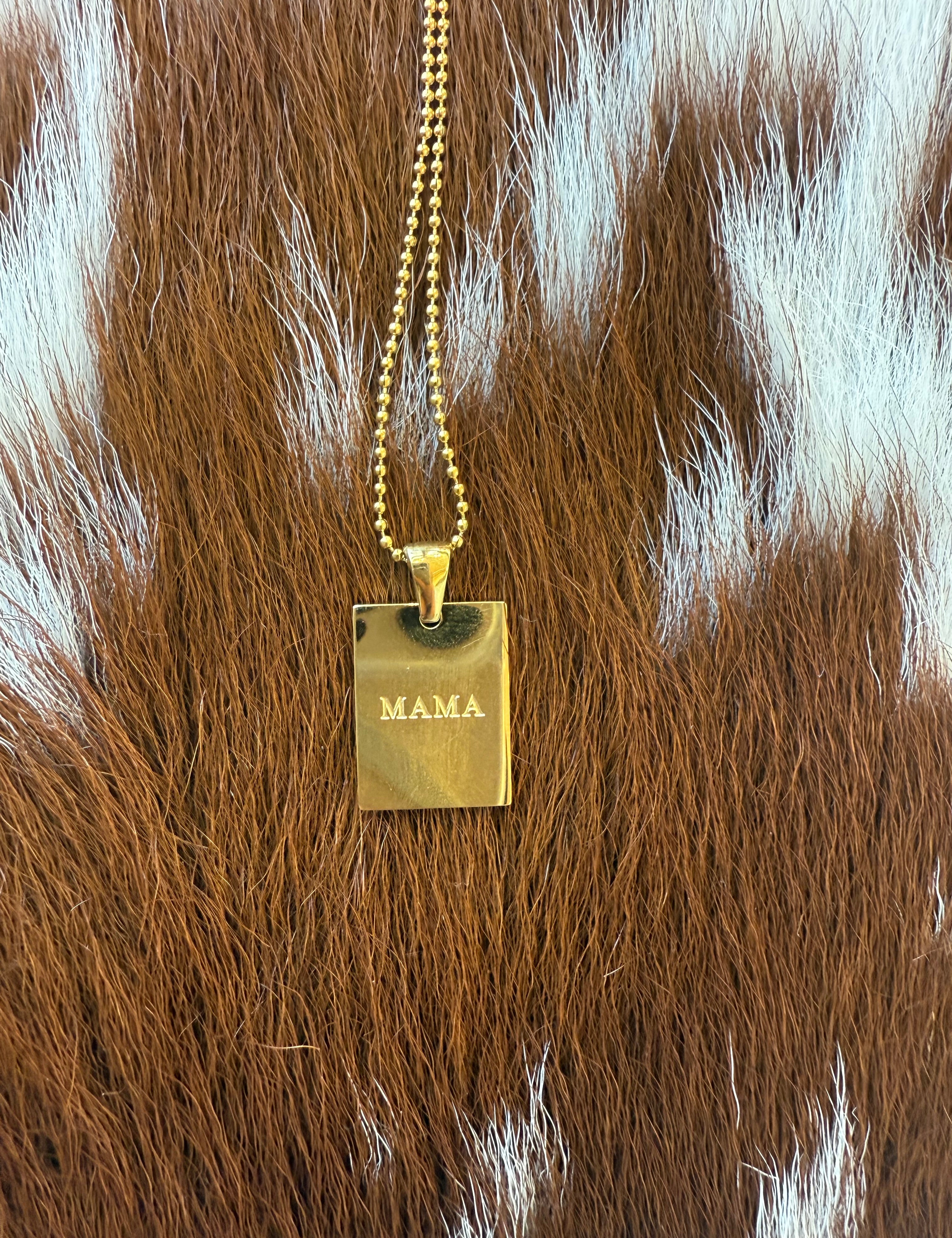  A gold rectangular pendant necklace with the word "MAMA" engraved on it, displayed against a textured brown and white cowhide background.