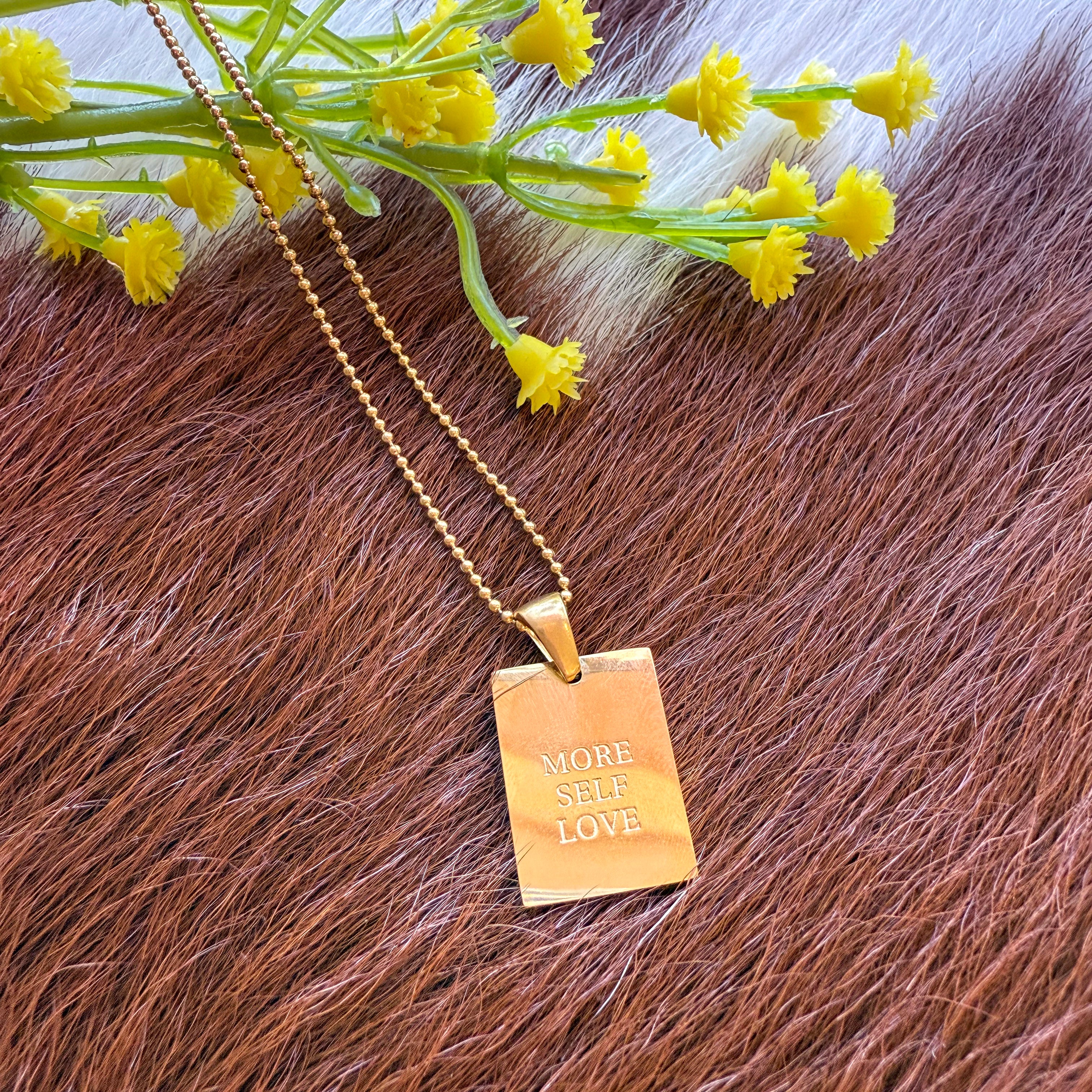  A gold rectangular pendant necklace engraved with the words "MORE SELF LOVE," displayed on a brown and white cowhide background with a small bunch of yellow flowers placed above the pendant.