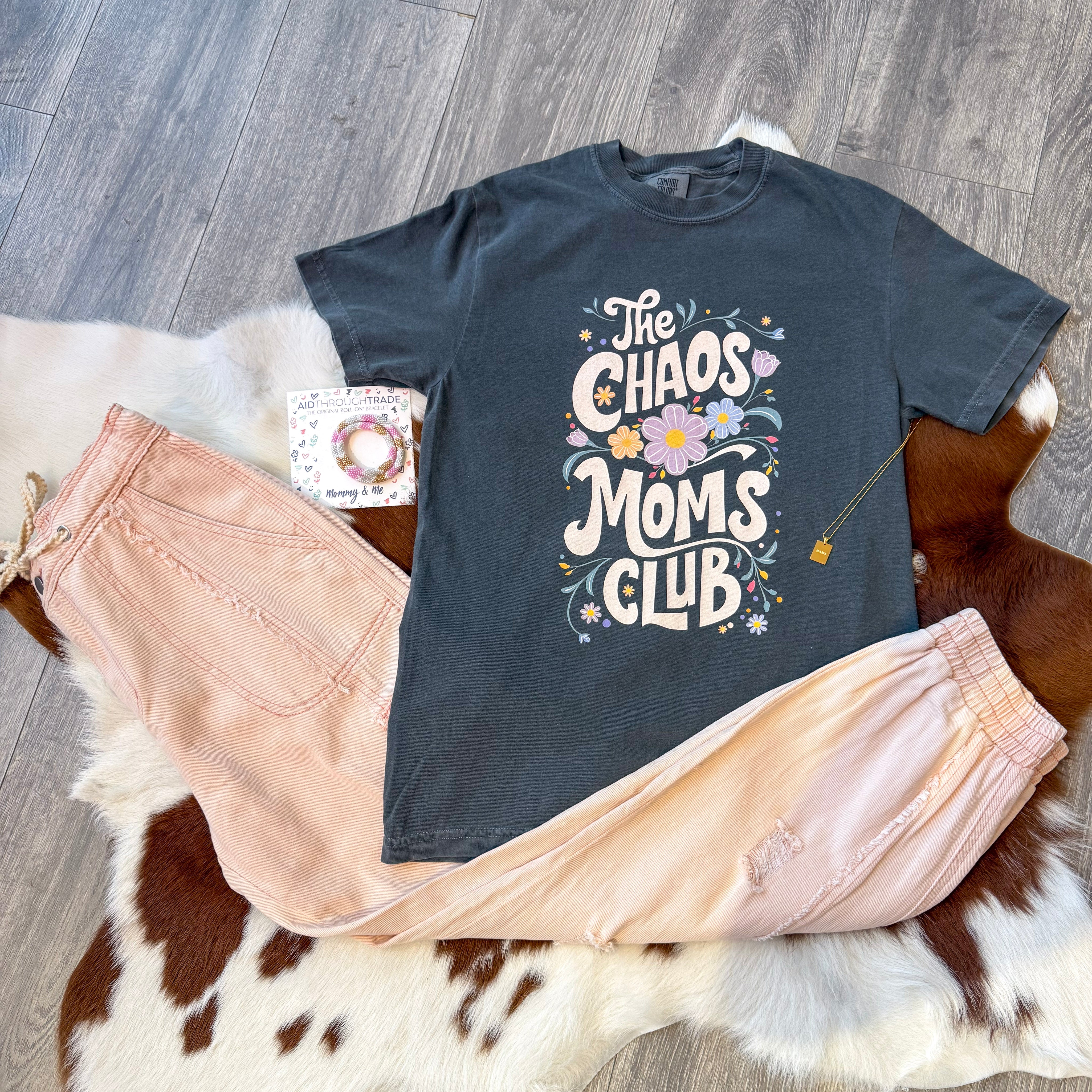 Flat lay of a boutique outfit featuring a dark gray “The Chaos Moms Club” graphic tee with playful floral lettering, styled with blush pink jogger pants, a set of colorful bracelets, and a gold padlock necklace, all laid out on a cowhide rug over wood flooring.