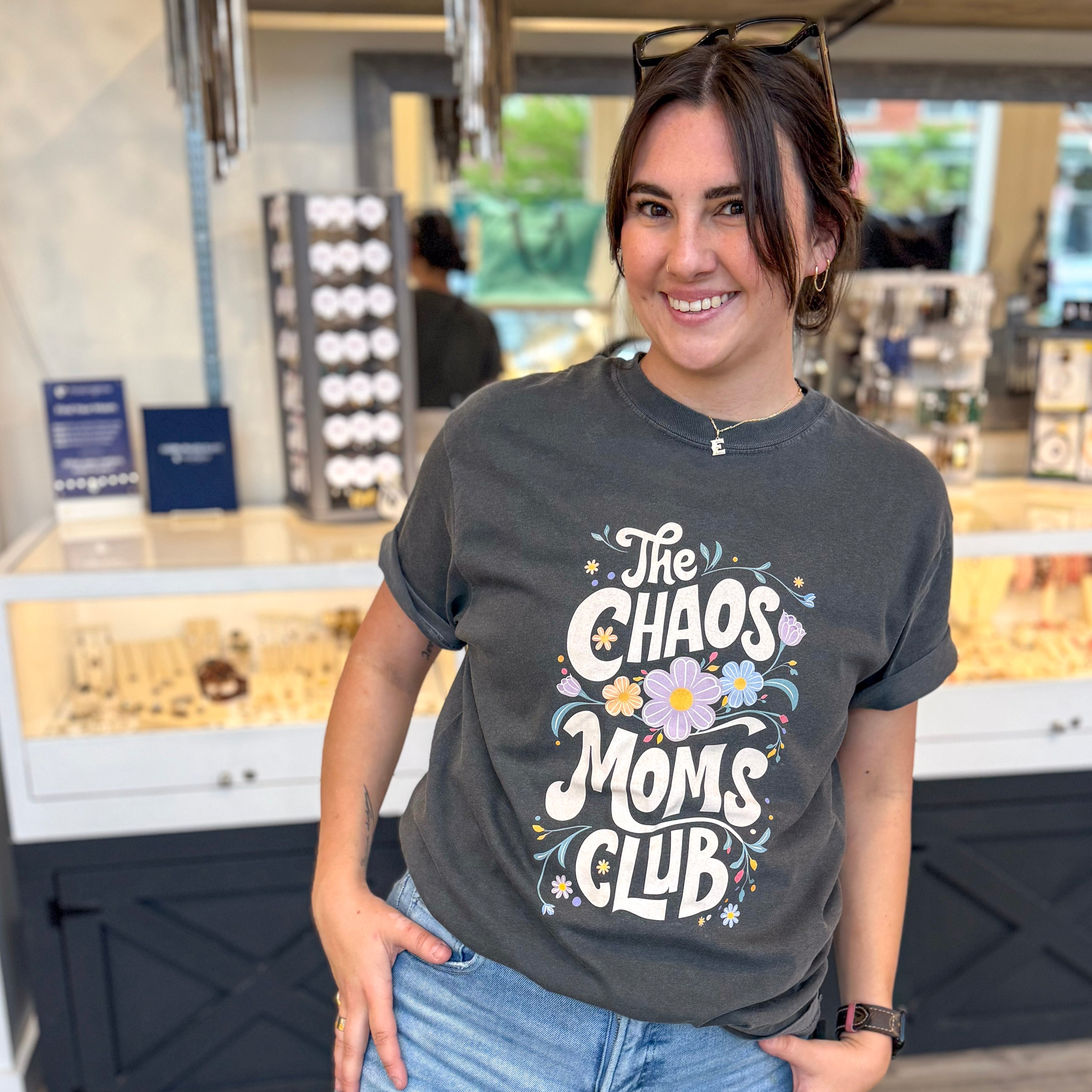 Smiling woman standing inside a boutique wearing a charcoal gray graphic tee that reads “The Chaos Moms Club” with colorful floral accents, styled with rolled sleeves, light wash jeans, hoop earrings, and sunglasses on her head, posed in front of a jewelry display case.