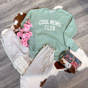 Flat lay of a cozy boutique outfit featuring a sage green sweatshirt with white “COOL MOMS CLUB” lettering, paired with light taupe jogger pants, a floral and cowhide wristlet bag, a white crochet top, and pink faux flowers, all displayed on a cowhide rug over wood flooring.