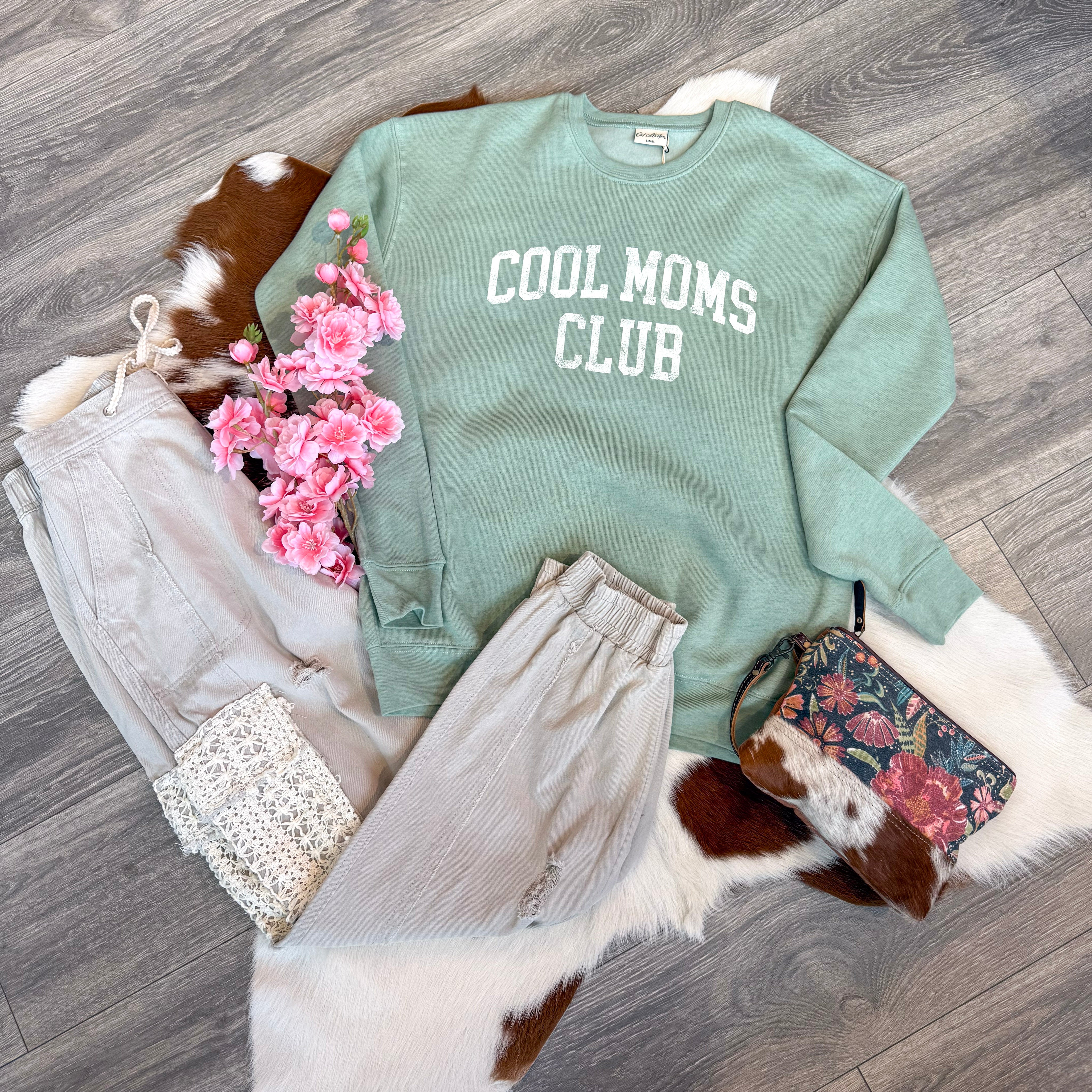 Flat lay of a cozy boutique outfit featuring a sage green sweatshirt with white “COOL MOMS CLUB” lettering, paired with light taupe jogger pants, a floral and cowhide wristlet bag, a white crochet top, and pink faux flowers, all displayed on a cowhide rug over wood flooring.