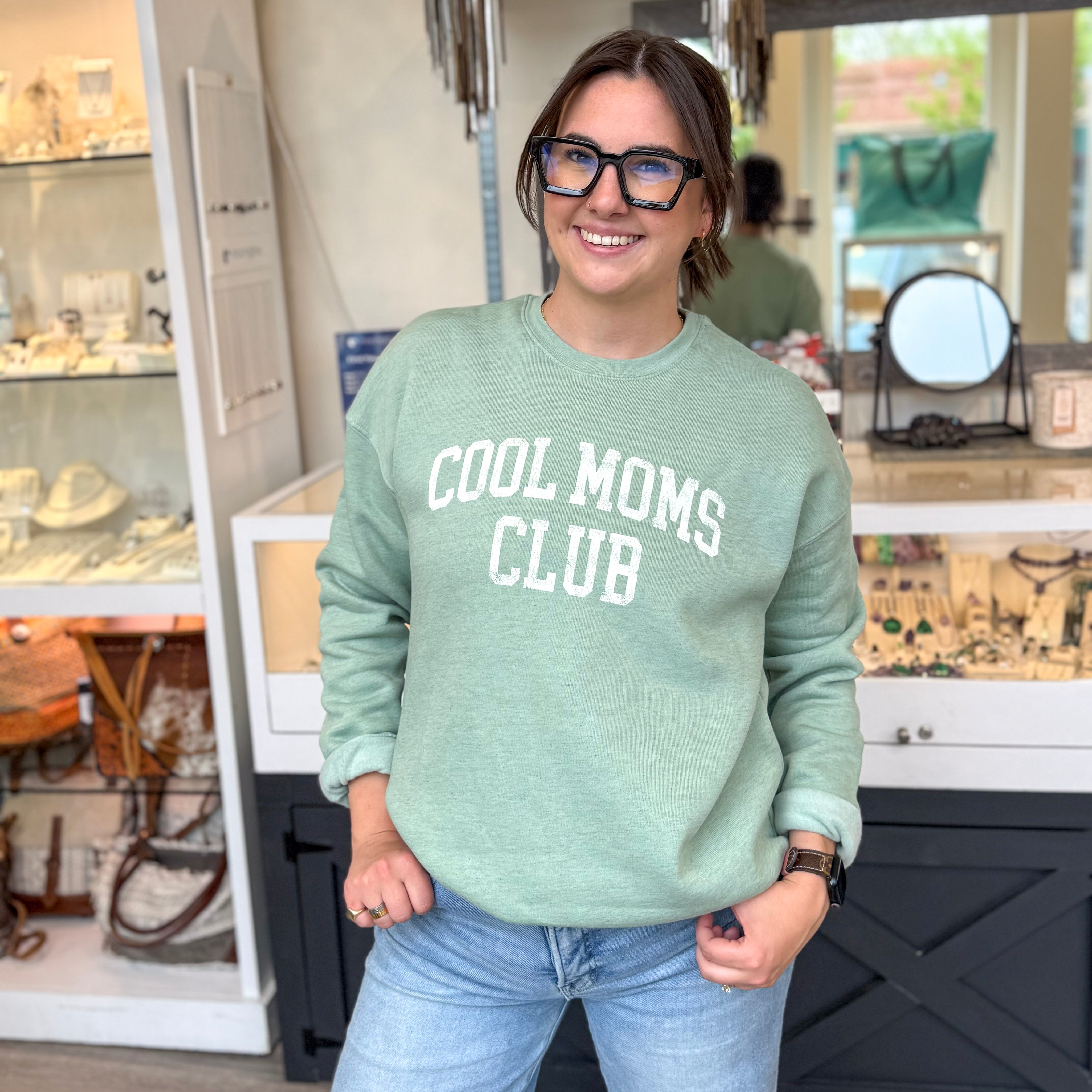 Woman smiling in a boutique wearing a light sage green sweatshirt with white block lettering that reads “COOL MOMS CLUB,” styled with light wash jeans, black-framed glasses, an Apple Watch, and gold jewelry, standing in front of a jewelry and accessories display.