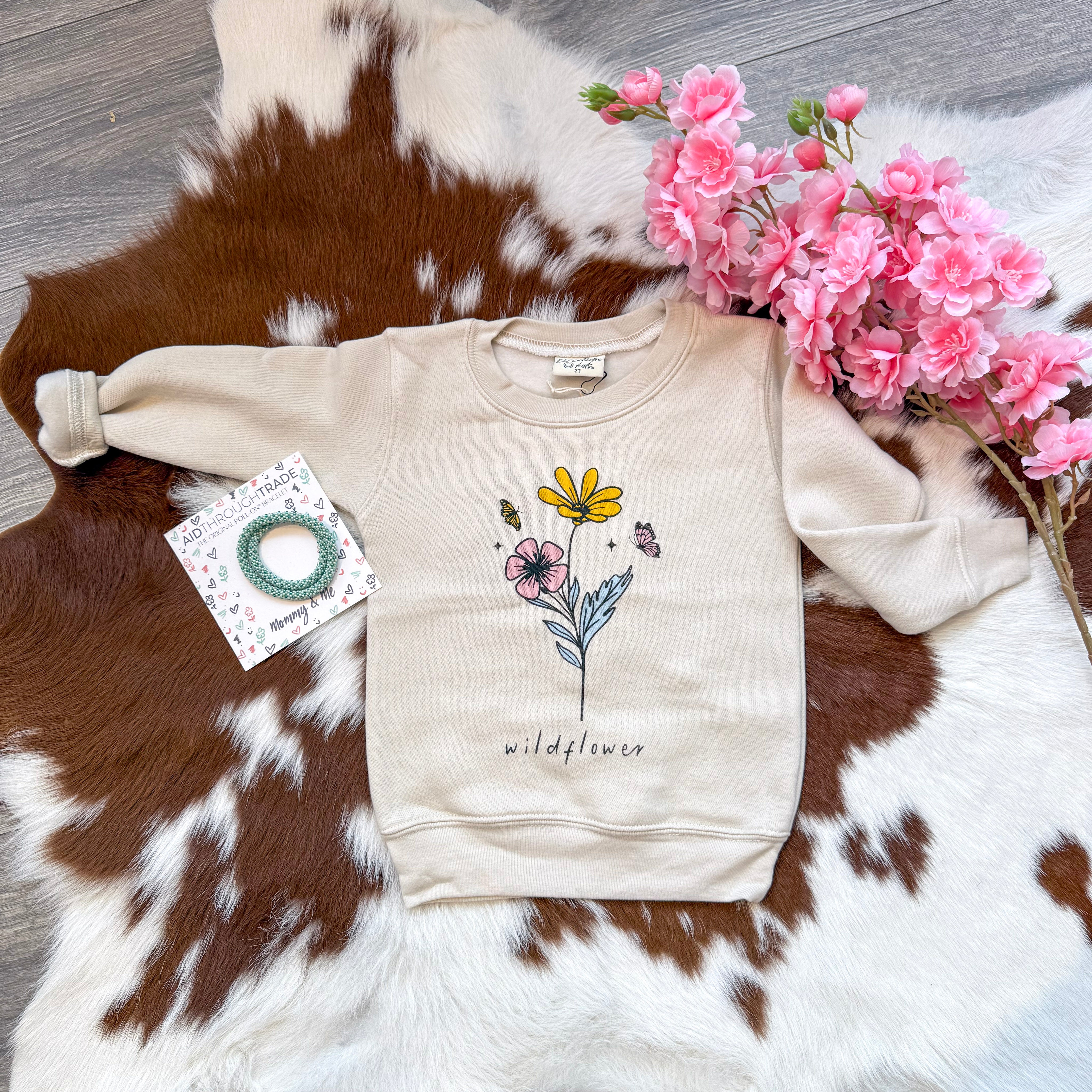 Flat lay of a cream toddler sweatshirt featuring a whimsical floral and butterfly graphic with the word “wildflower” underneath, styled with a mint green “Mommy & Me” bracelet set and pink faux flowers, all arranged on a brown and white cowhide rug over wood flooring.