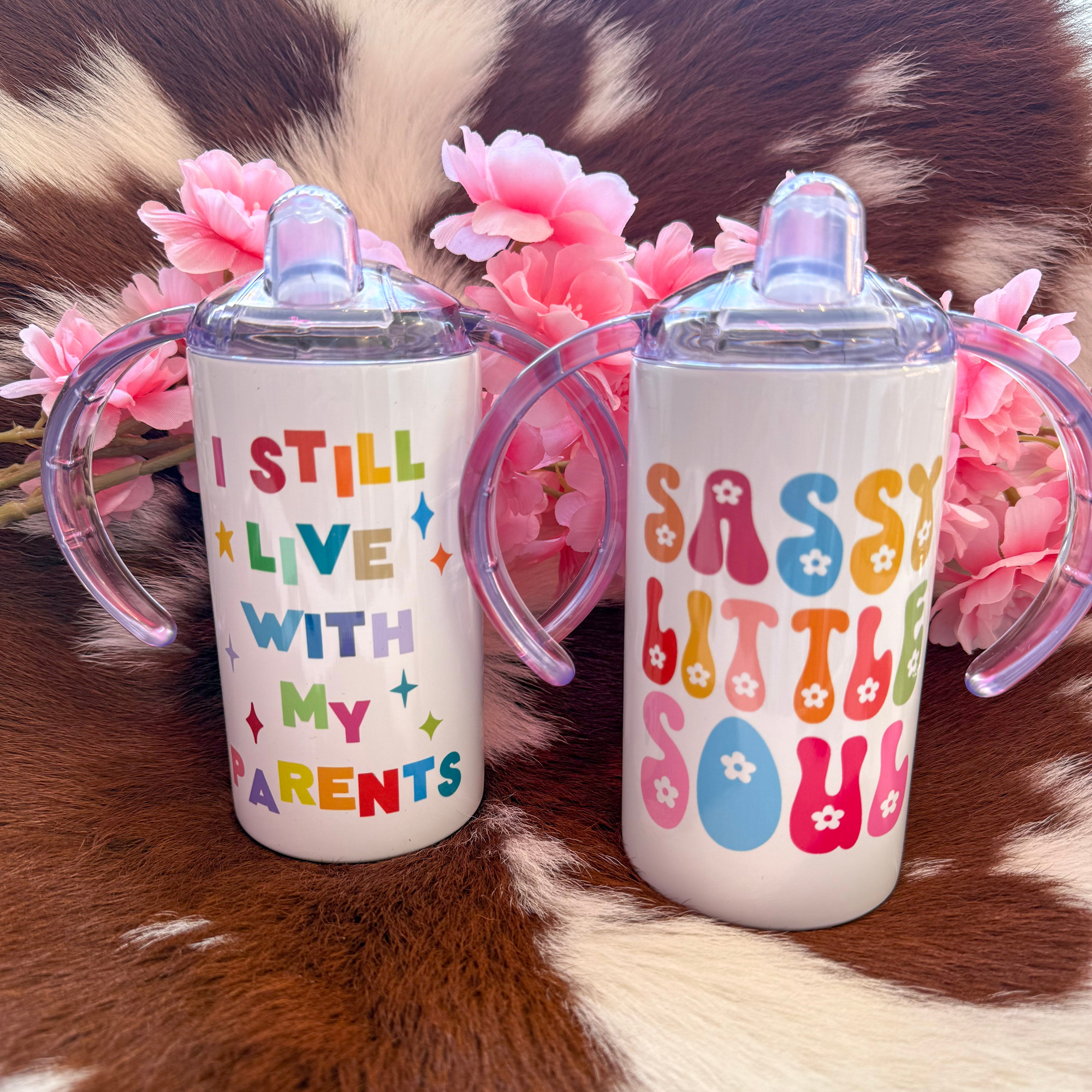 Two colorful toddler sippy cups with funny phrases. One says “I Still Live With My Parents” in rainbow letters, and the other says “Sassy Little Soul” in retro bubble letters, both surrounded by pink flowers on a cowhide background.