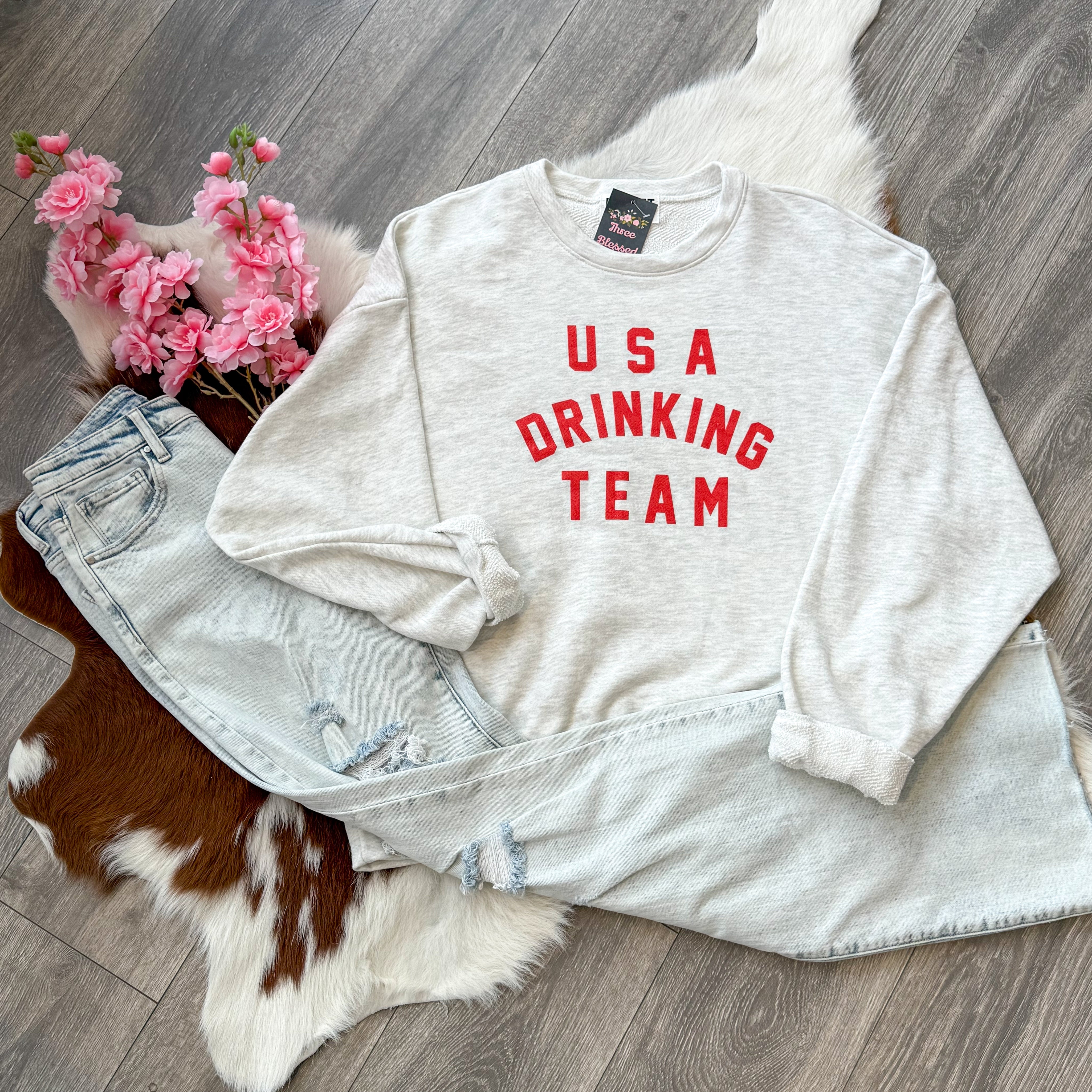 Flat lay of a light gray sweatshirt with bold red "USA Drinking Team" text, styled with light-wash distressed jeans, pink flowers, and a faux cowhide rug on a wood floor for a festive, casual look.