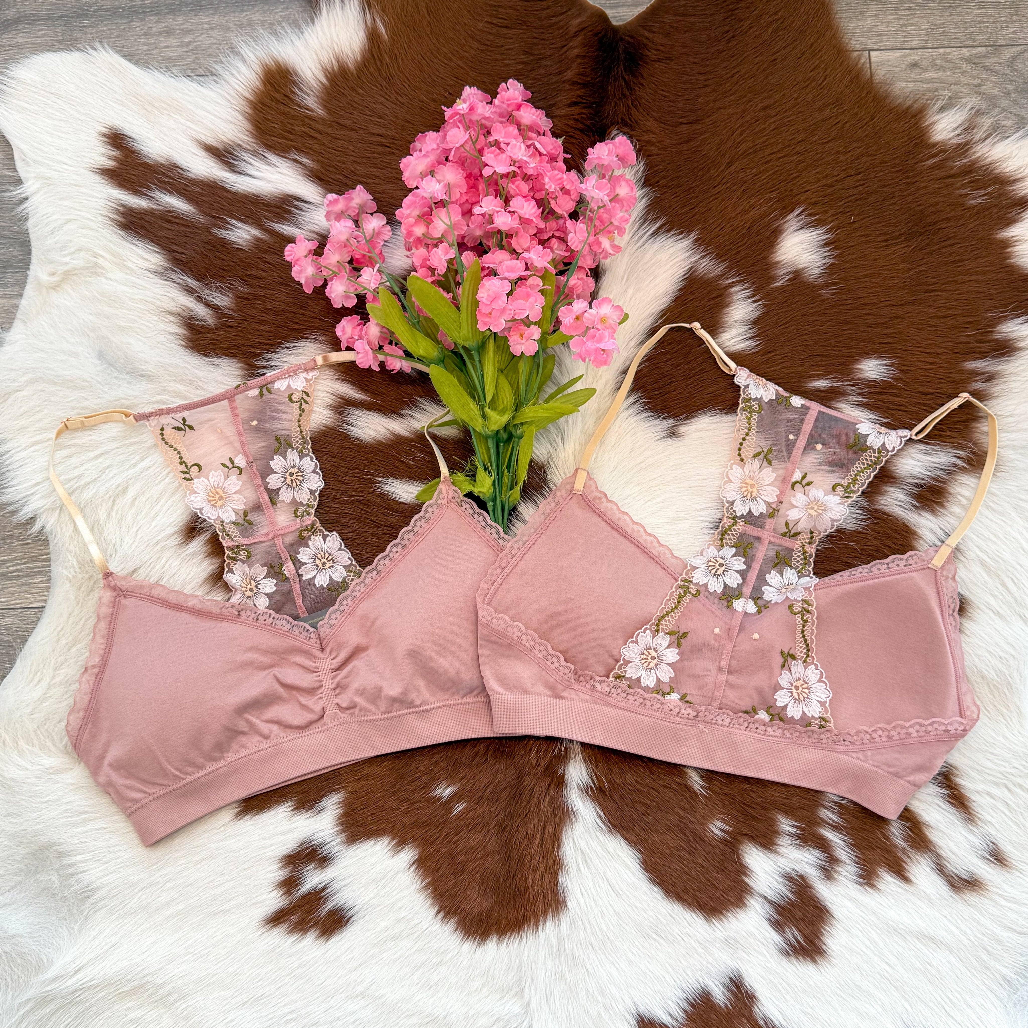 Dusty rose bralette featuring a sheer mesh panel with delicate white floral embroidery, laid on a brown and white cowhide rug with a bunch of vibrant pink faux flowers placed in the center for a soft and romantic vibe.