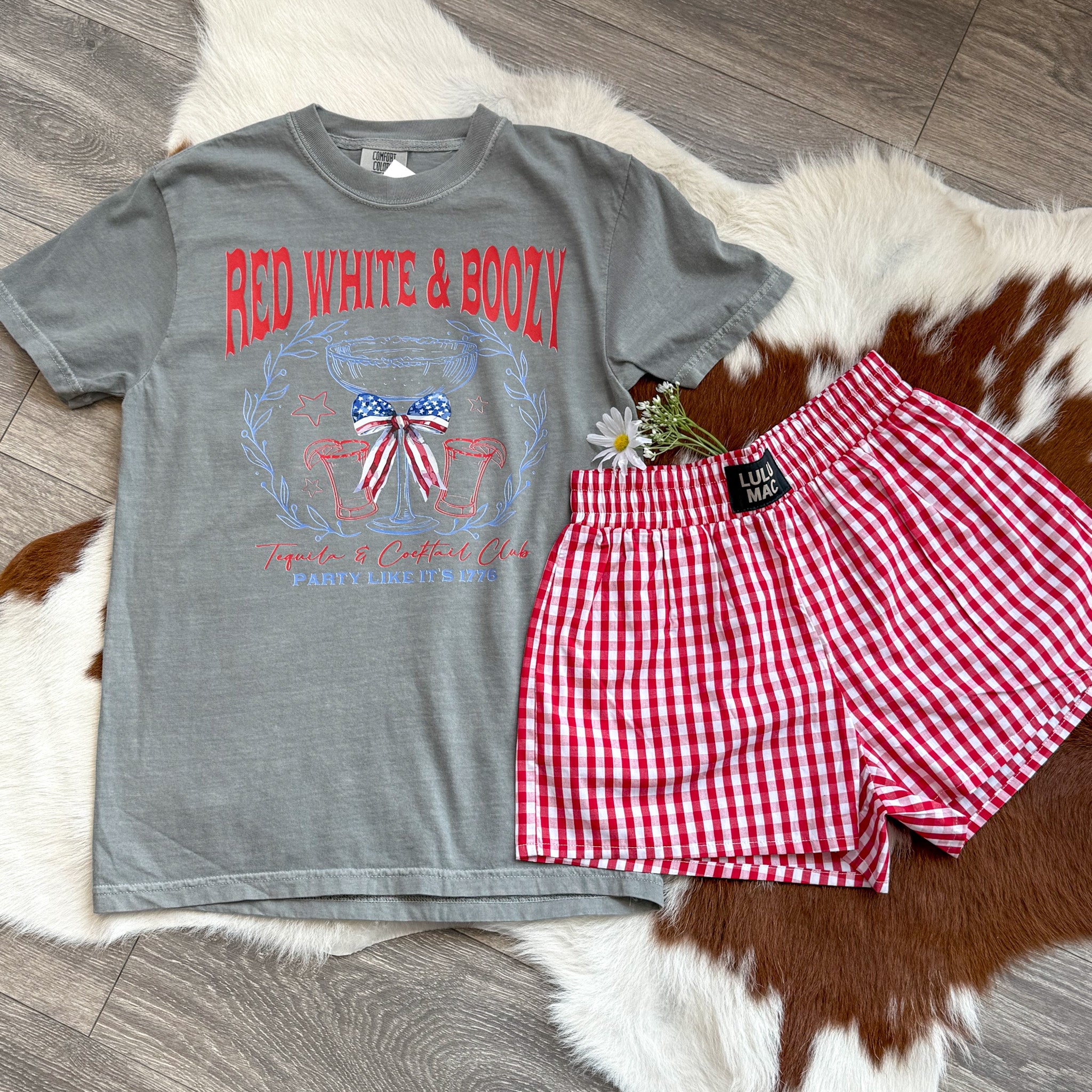 Flat lay of a grey graphic t-shirt reading “Red White & Boozy” with patriotic accents and cocktail-themed artwork, styled for the Fourth of July.

