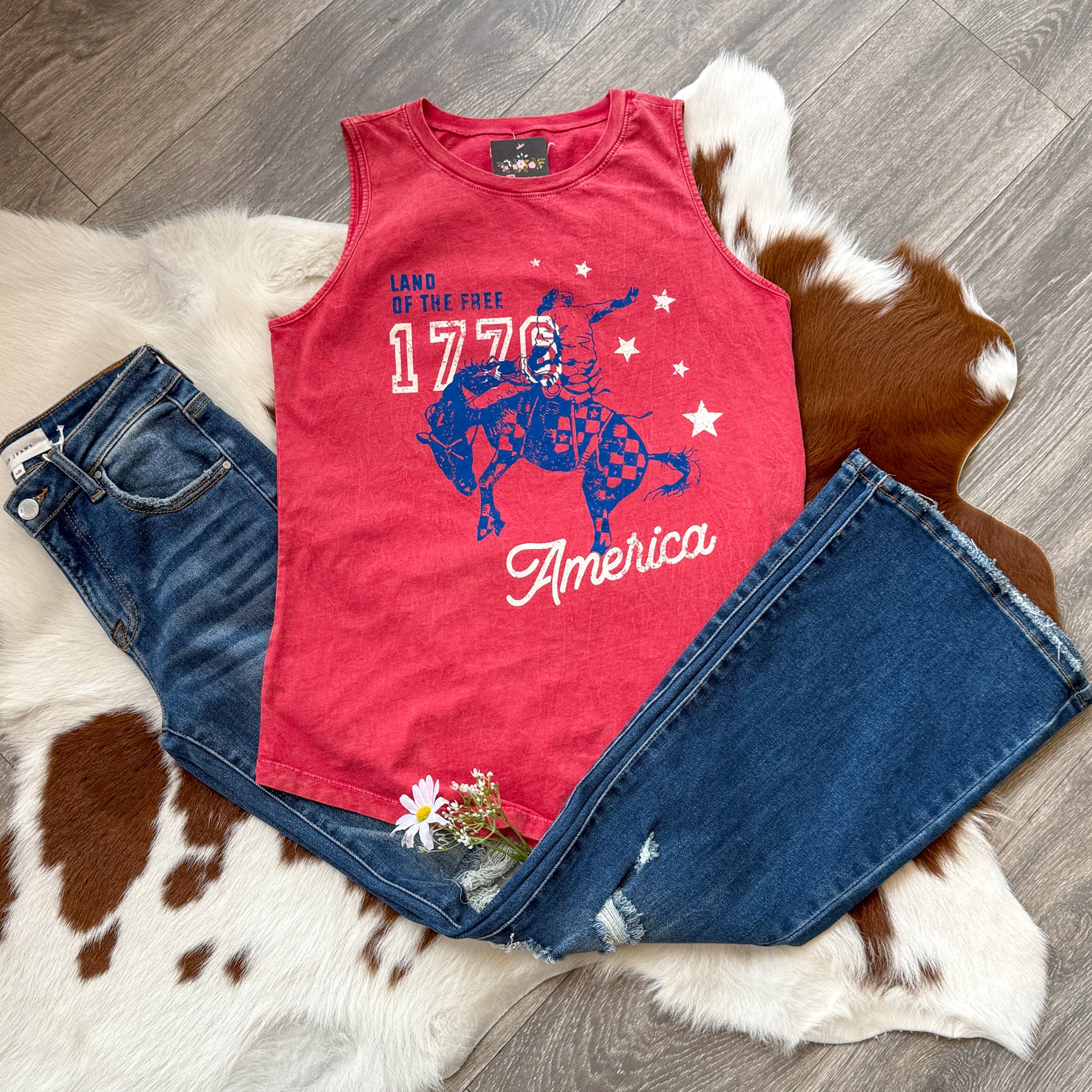 Flat lay of a red sleeveless tank top with a graphic that reads “Land of the Free 1776 America,” featuring a rider on a bucking horse in blue and red patterns, paired with distressed denim jeans and styled on a cowhide rug with white daisies tucked in the pocket—perfect for a patriotic and western-inspired 4th of July outfit.
