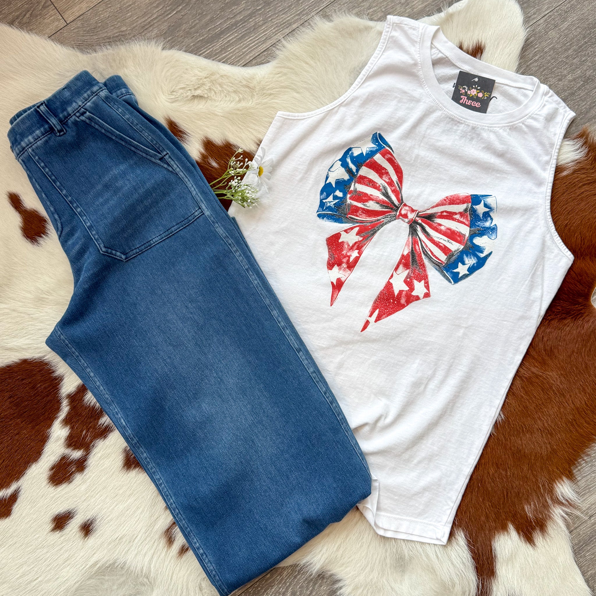 Flat lay of a white sleeveless tank top featuring a large red, white, and blue bow graphic with stars and stripes, paired with high-waisted blue jeans. The outfit is styled on a cowhide rug with white daisies tucked in the waistband—perfect for a cute and festive Fourth of July look.