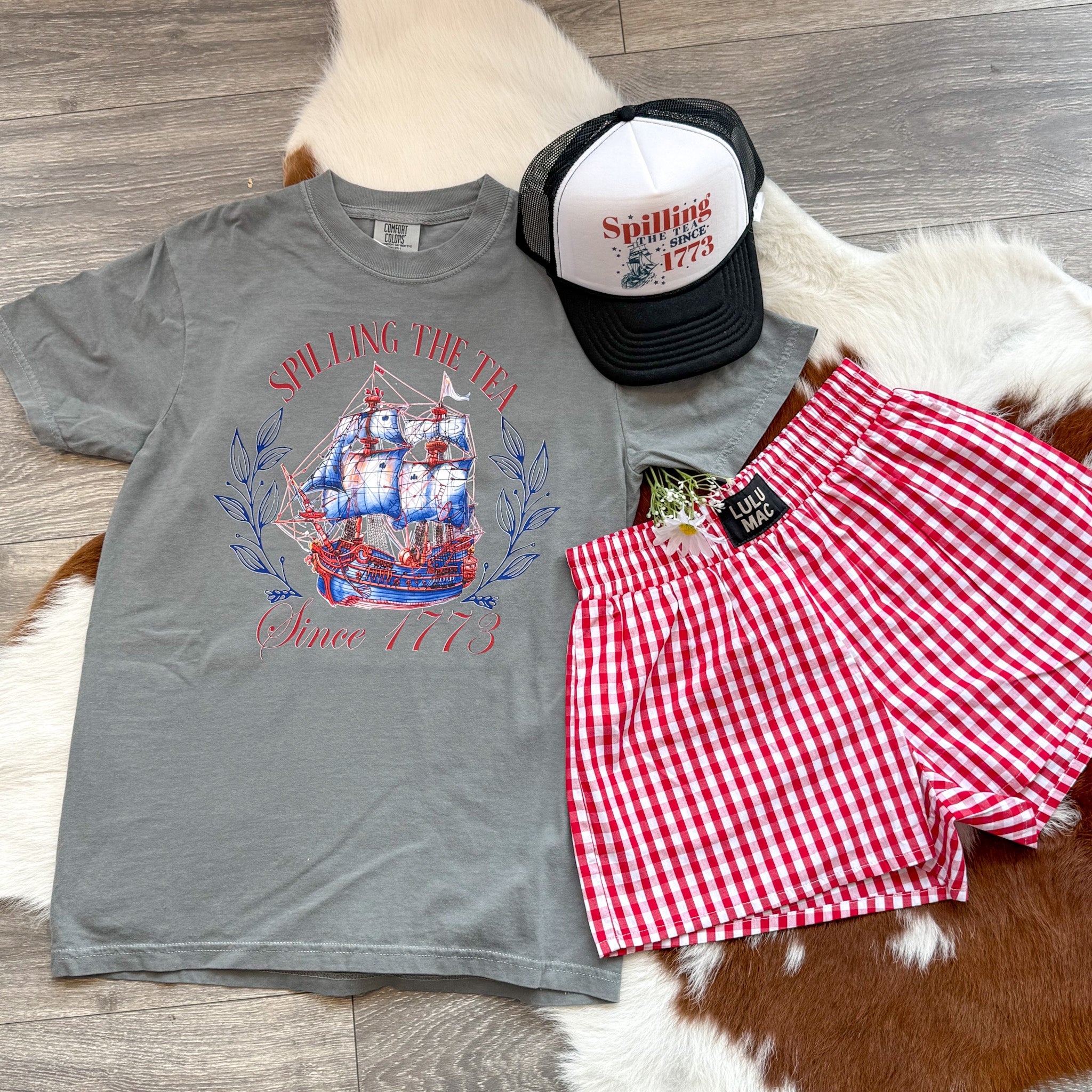 Flat lay of a grey graphic tee with a colonial ship and the phrase “Spilling the Tea Since 1773,” styled with red gingham shorts and a matching trucker hat on a cowhide rug—perfect for a cheeky and patriotic Fourth of July outfit.