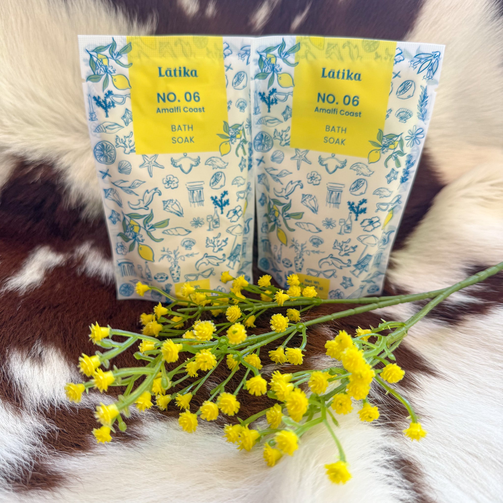 Two packets of Latika No. 06 Amalfi Coast Bath Soak with a blue and yellow lemon-themed design, styled on a brown-and-white cowhide rug with yellow flowers in the foreground.