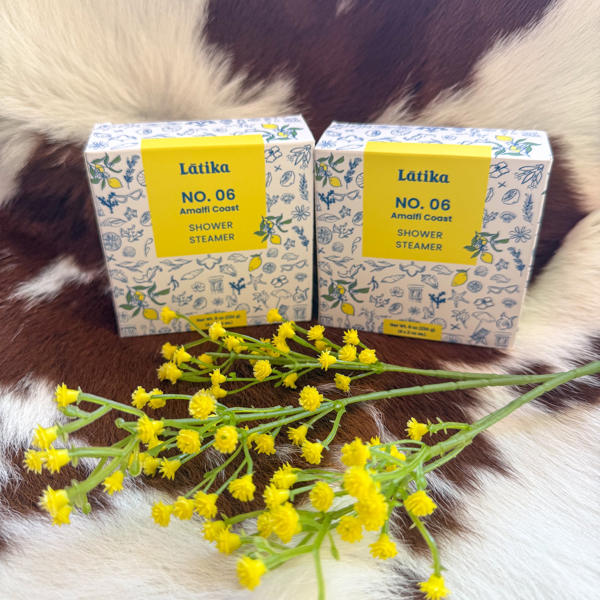 "Two Latika Amalfi Coast No. 06 Shower Steamer boxes with floral blue illustrations and bright yellow accents, styled on cowhide with fresh yellow flowers"