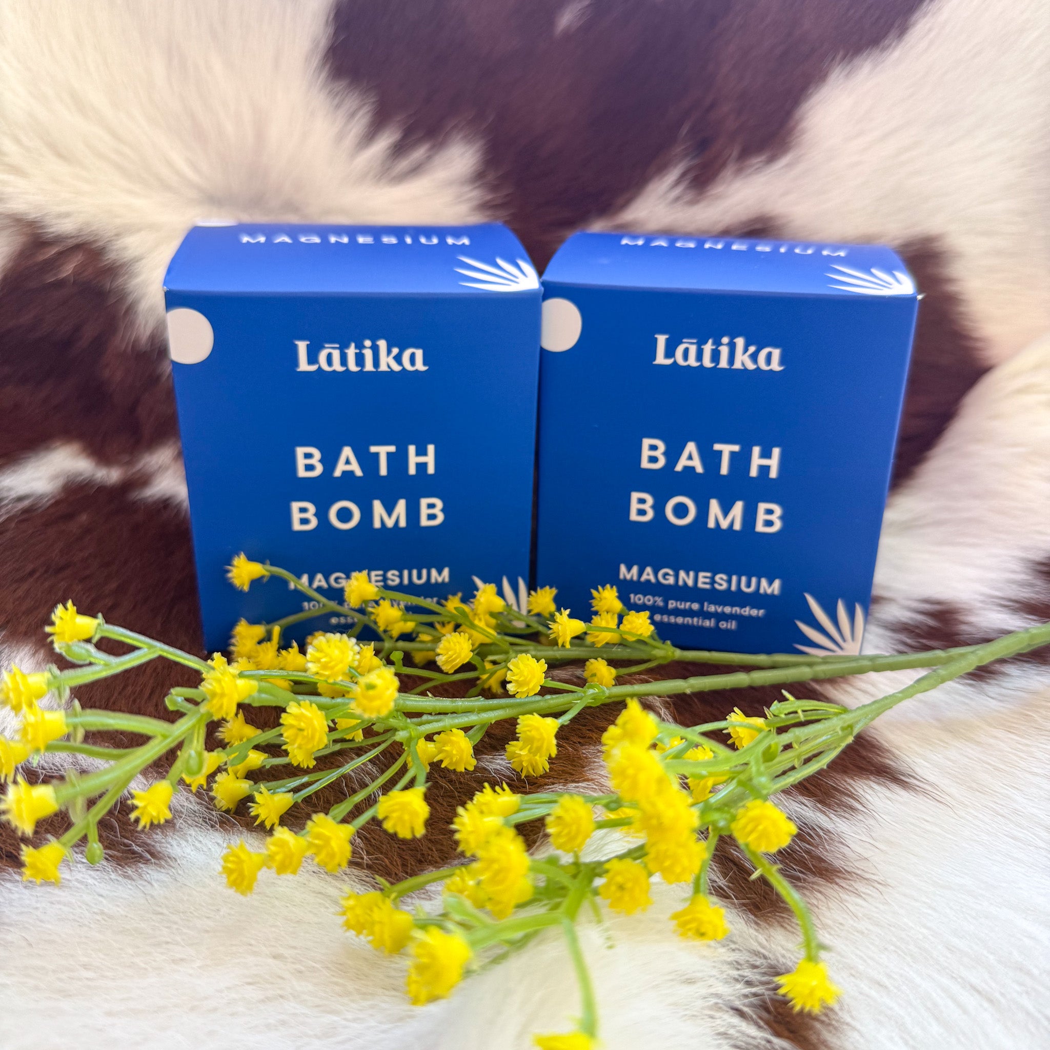 Two blue boxes of Latika Magnesium Bath Bombs with white text, featuring lavender essential oil, displayed on a brown-and-white cowhide rug with small yellow flowers in the foreground.