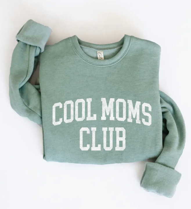 Sage green fleece crewneck sweatshirt folded with white "Cool Moms Club" text printed in bold varsity letters.