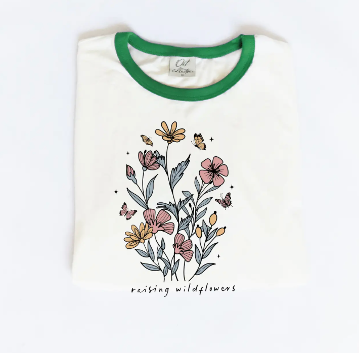A cream-colored graphic tee with a green collar featuring a floral print with butterflies and the text "raising wildflowers" written underneath.