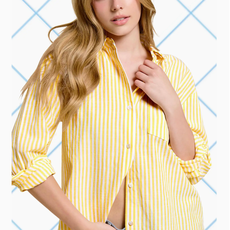 A woman wearing a yellow and white striped button-up shirt, smiling softly at the camera. The shirt features a relaxed fit and is partially unbuttoned, styled with rolled-up sleeves. She is paired with denim shorts, showcasing a casual and chic look against a light blue geometric background.