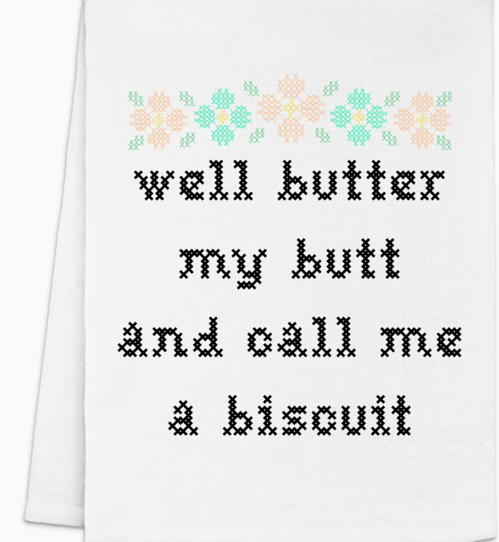  A white towel featuring the phrase "Well Butter My Butt and Call Me a Biscuit" in black cross-stitch font, adorned with colorful floral designs above.