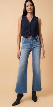 Woman wearing high-rise flare leg jeans in a medium blue wash, styled with a navy vest and black heeled boots against a tan background.