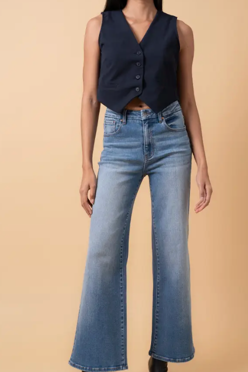 Woman wearing high-rise flare leg jeans in a medium blue wash, styled with a navy vest and black heeled boots against a tan background.