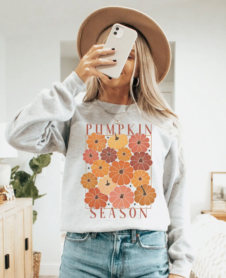 Image of a woman wearing a gray sweatshirt with a colorful pumpkin design, capturing the essence of fall fashion, while holding a phone and smiling.
