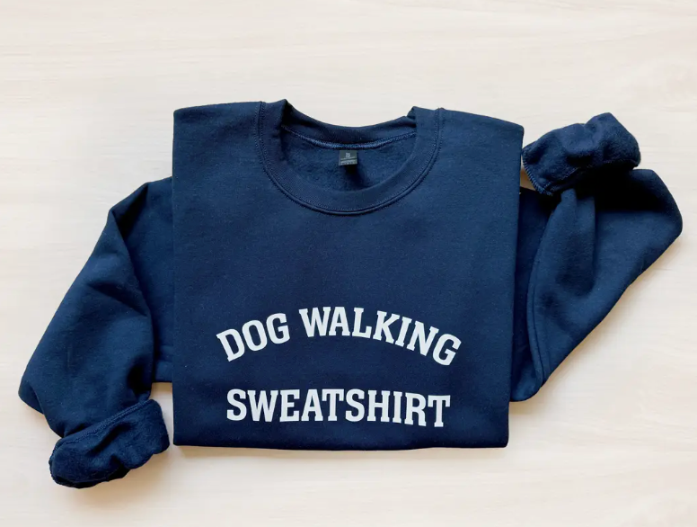 Close-up image of a navy sweatshirt with the text 'Dog Walking Sweatshirt' displayed prominently on the front, laid out on a light wooden surface.