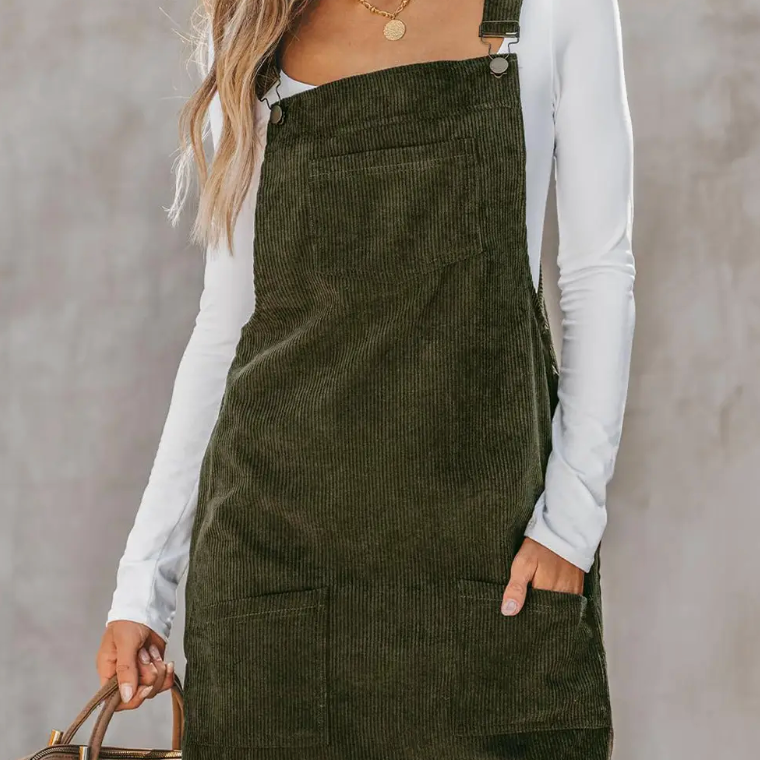 Model wearing an olive green corduroy overall dress layered over a white long-sleeve top, showcasing a stylish and casual fall look.